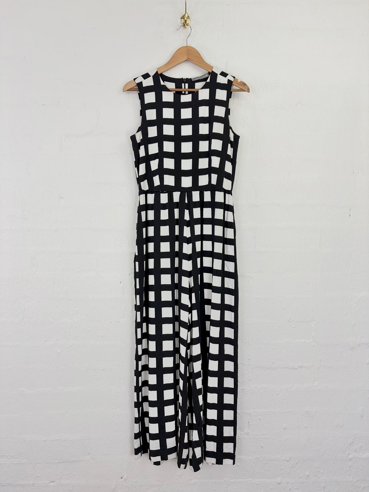 A sleeveless black and white checkered maxi dress hanging on a wooden hanger against a white painted brick wall.