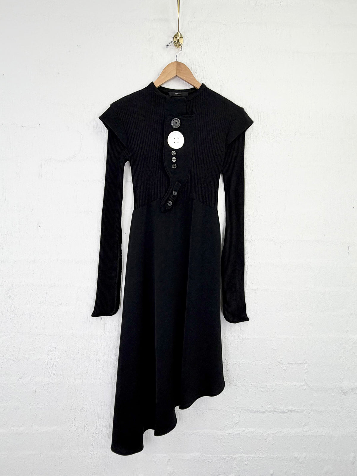 A black long-sleeve midi dress with a high-low asymmetrical hem and a buttoned neckline, hanging on a wooden hanger against a white painted brick wall.