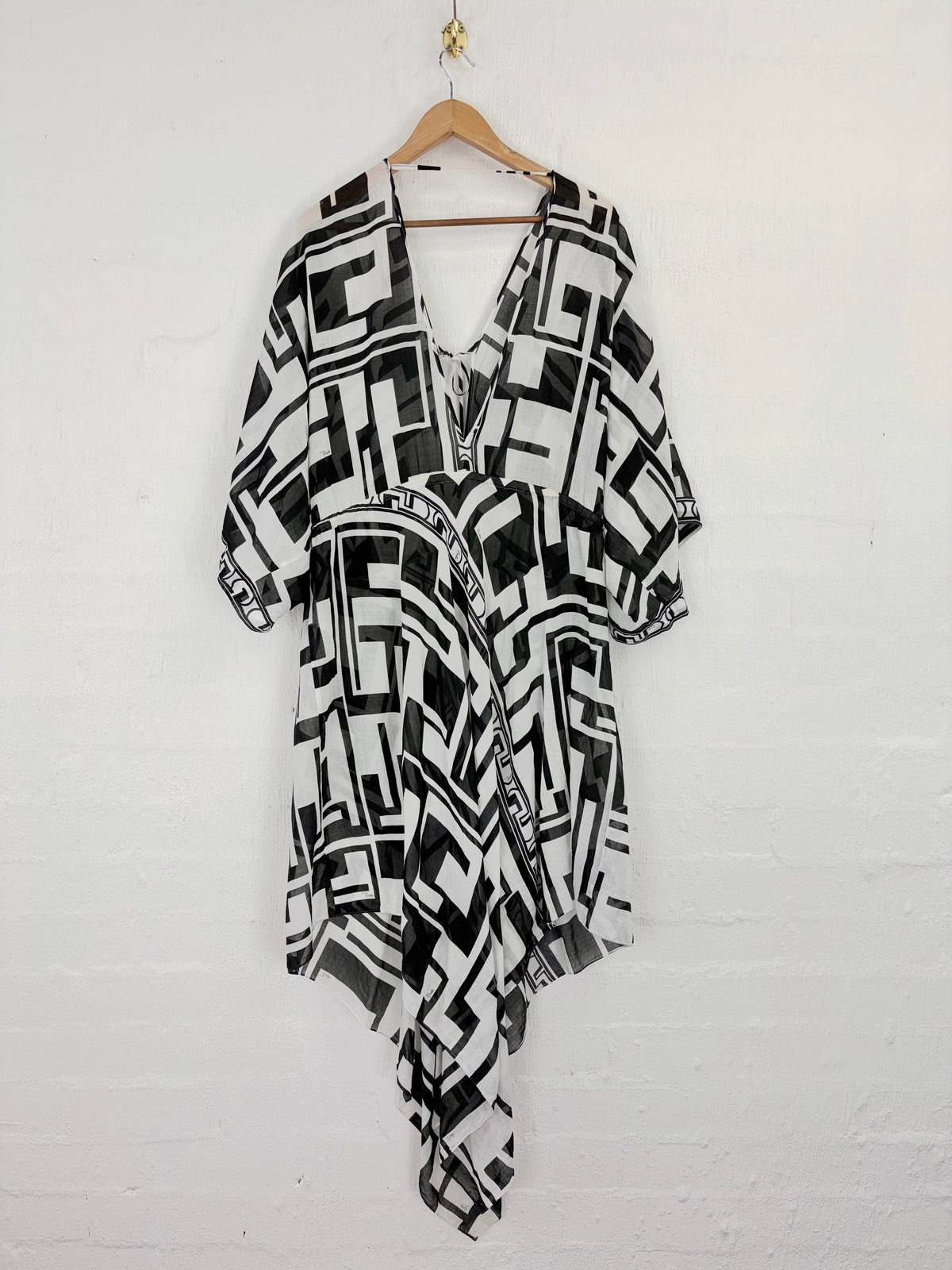A black and white geometric patterned wrap dress or kaftan hanging on a wooden hanger against a white painted brick wall.