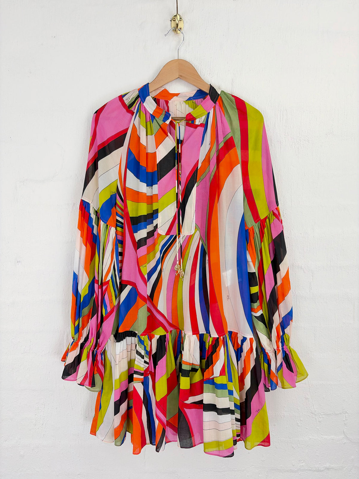 A long-sleeve, tiered blouse with a vibrant multicolor abstract stripe and swirl pattern, hanging on a wooden hanger against a white painted brick wall.