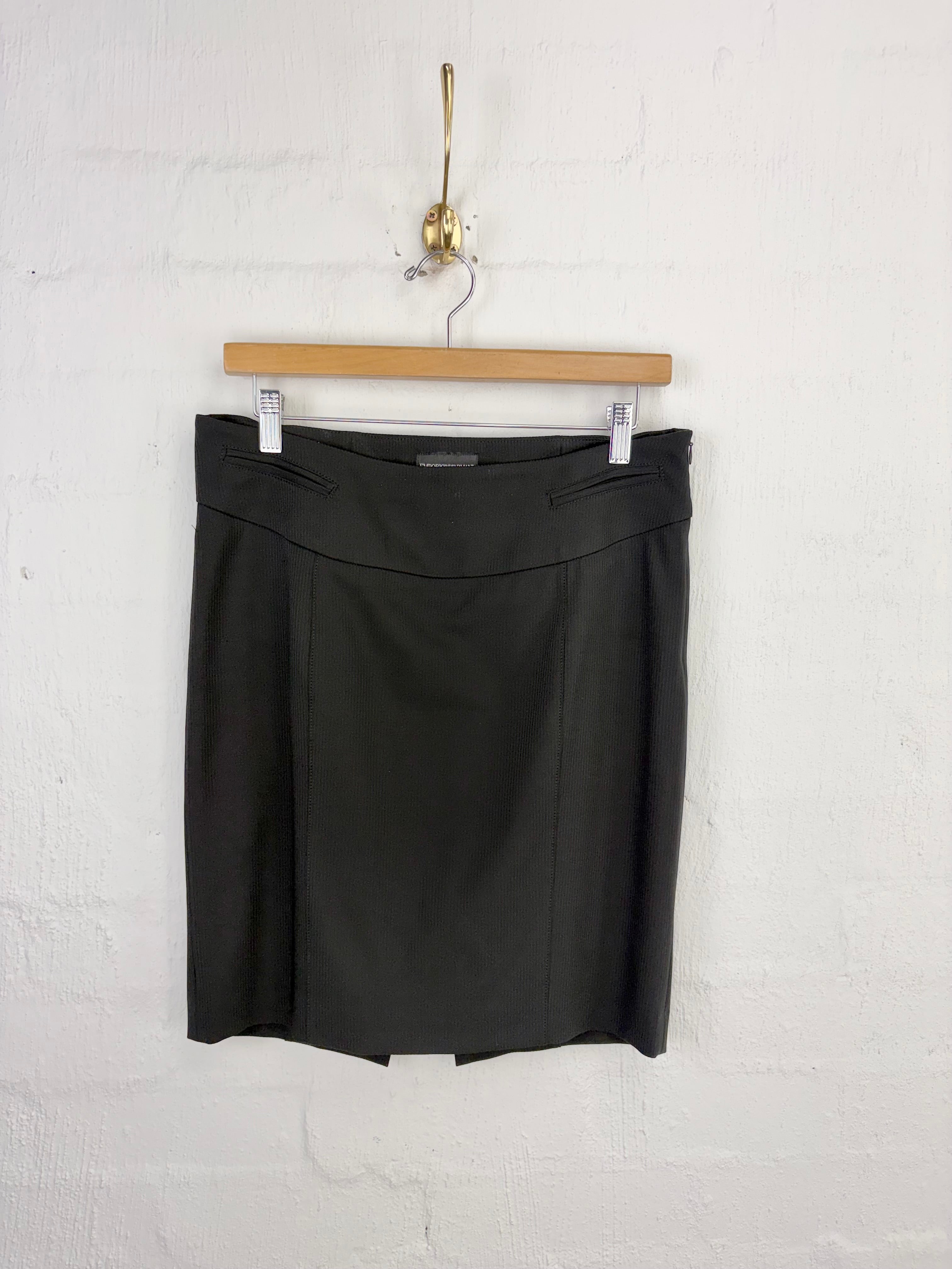A black mini skirt with a structured waistband and front pleats, hanging from a wooden hanger on a white painted brick wall.