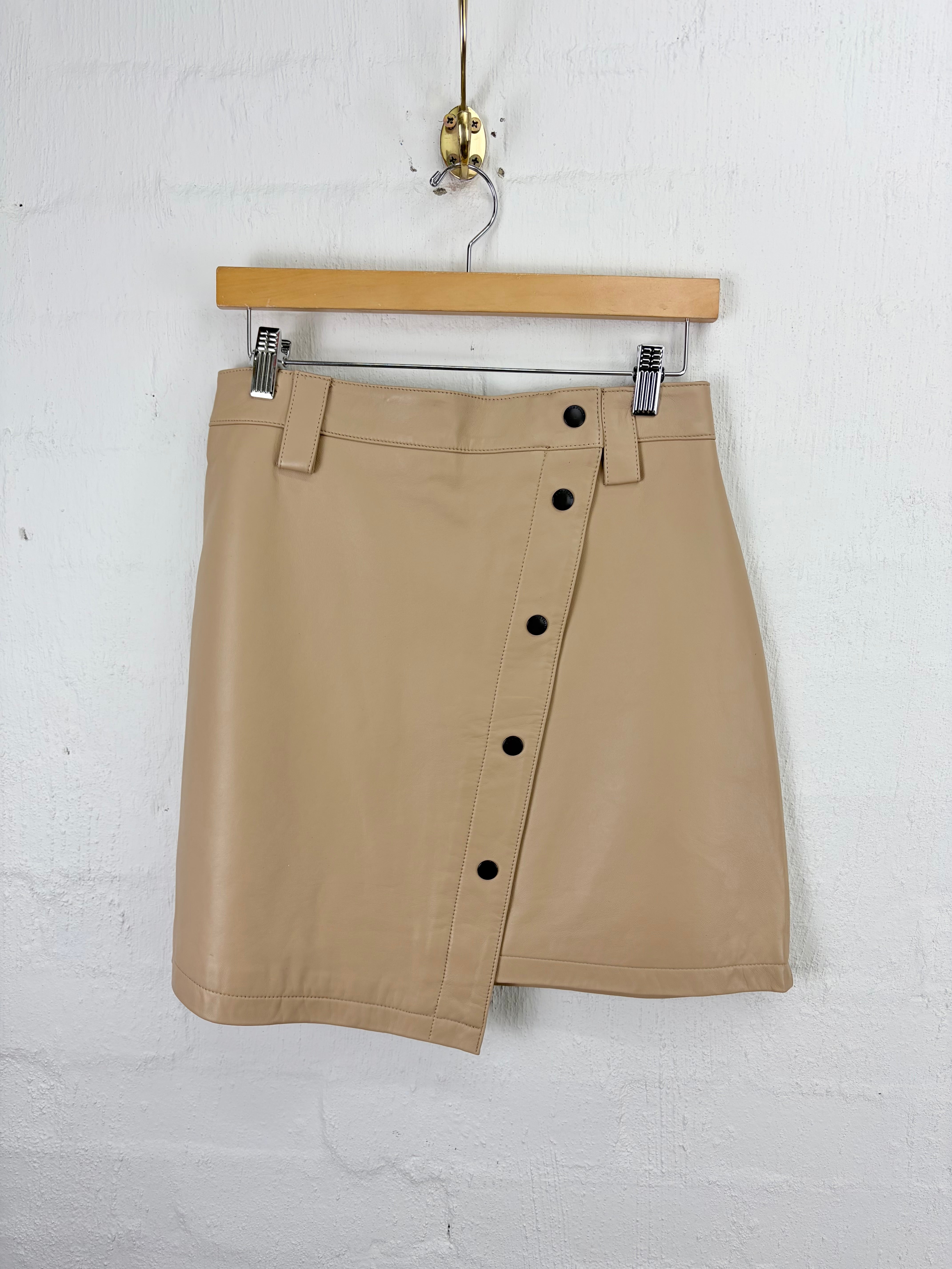 A beige leather mini skirt with an asymmetrical button-up front, hanging from a wooden hanger against a white painted brick wall.