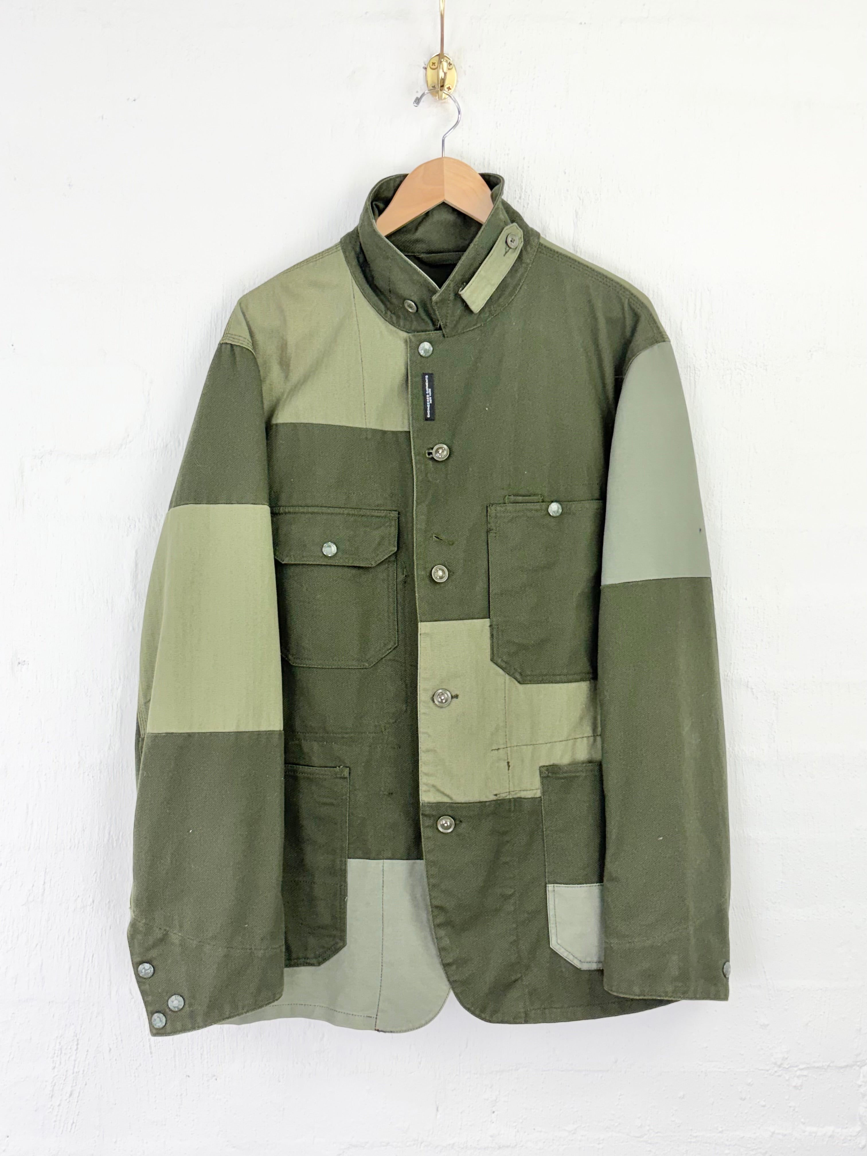 A vintage patchwork utility jacket in olive green and light khaki, hanging on a wooden hanger against a white painted brick wall.