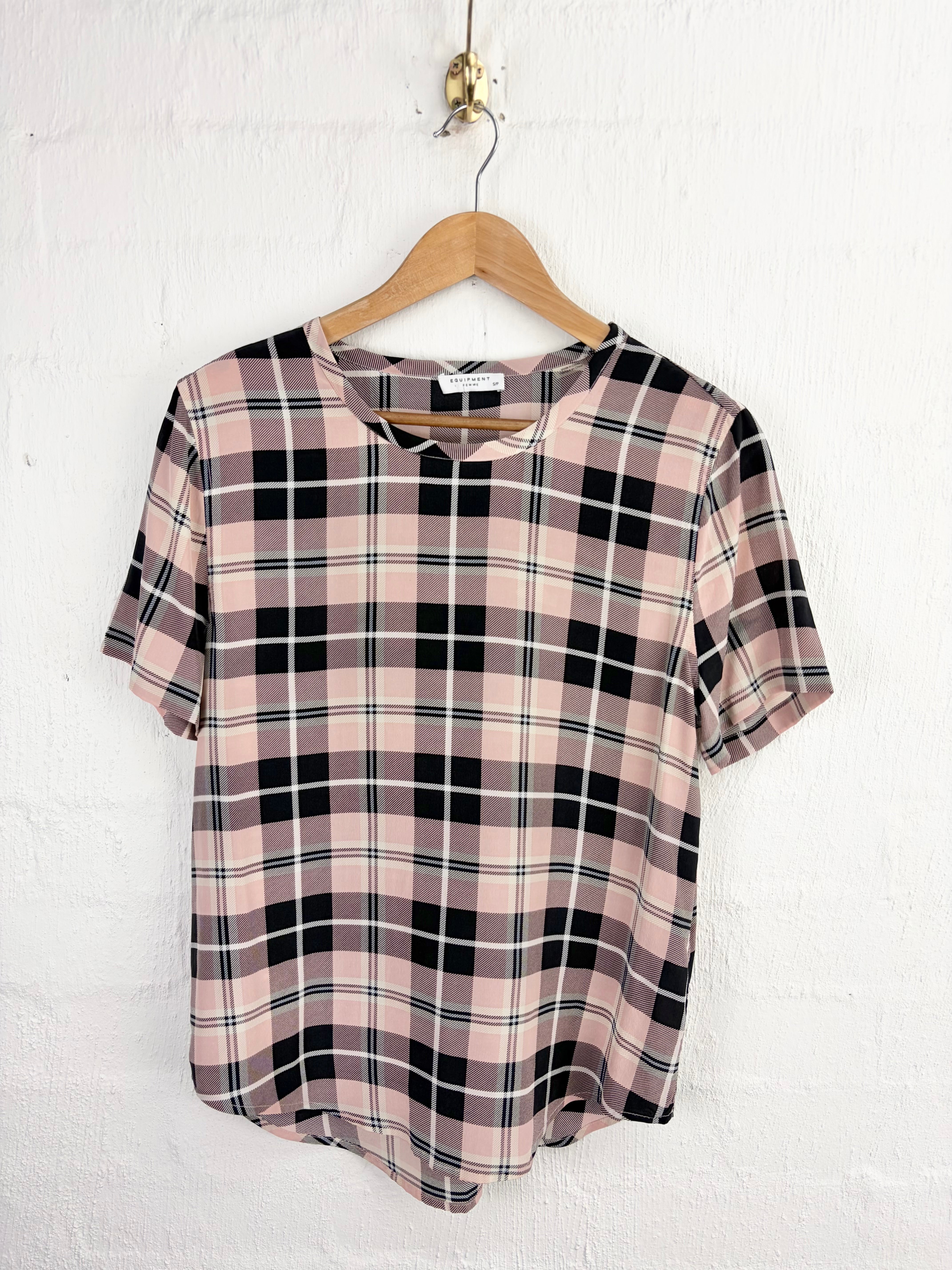 A short-sleeve plaid top in dusty pink, black, and white hanging on a wooden hanger against a white textured wall.