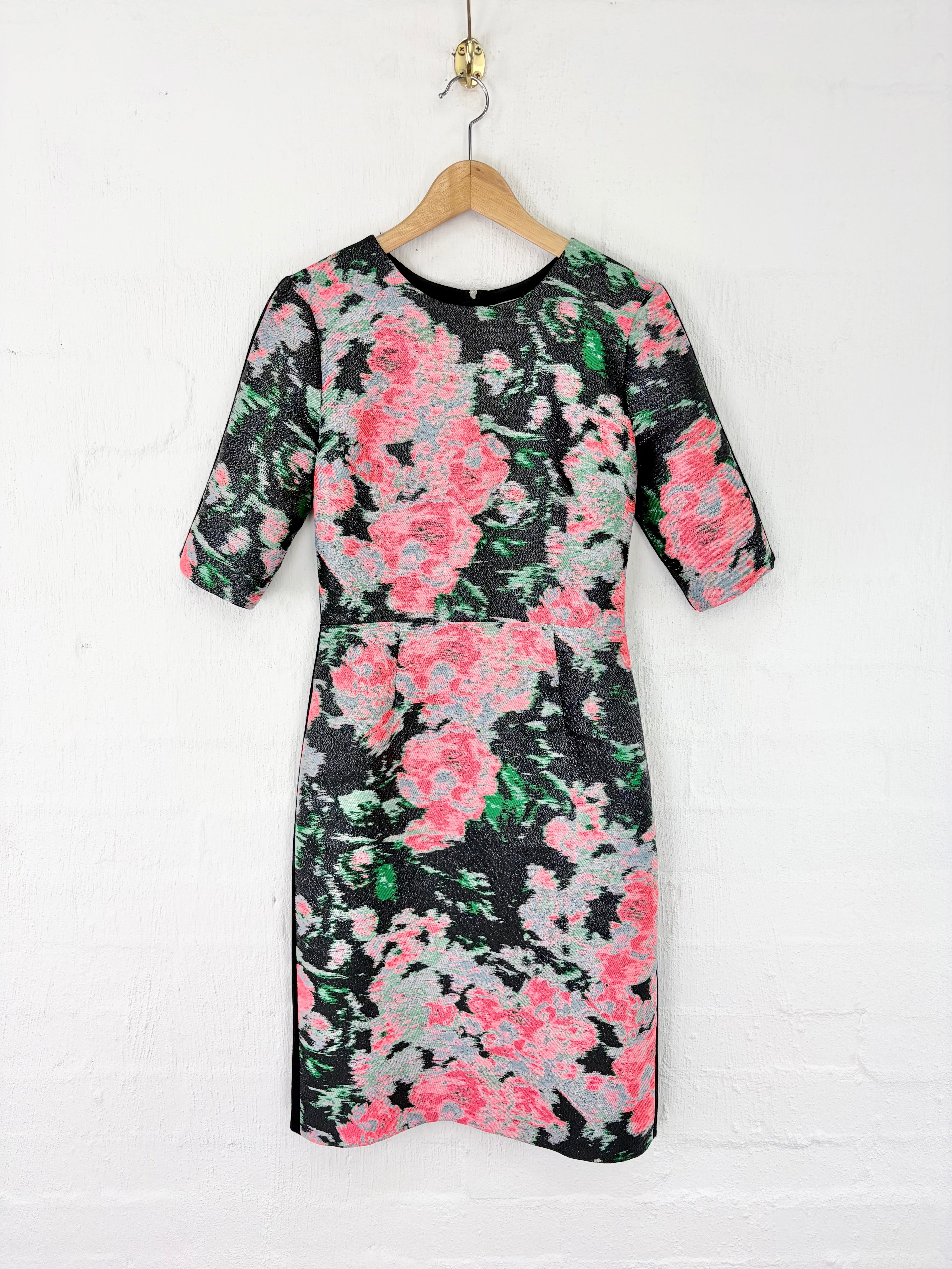 A knee-length black dress with a vibrant pink, green, and gray floral pattern, hanging on a wooden hanger against a white painted brick wall.