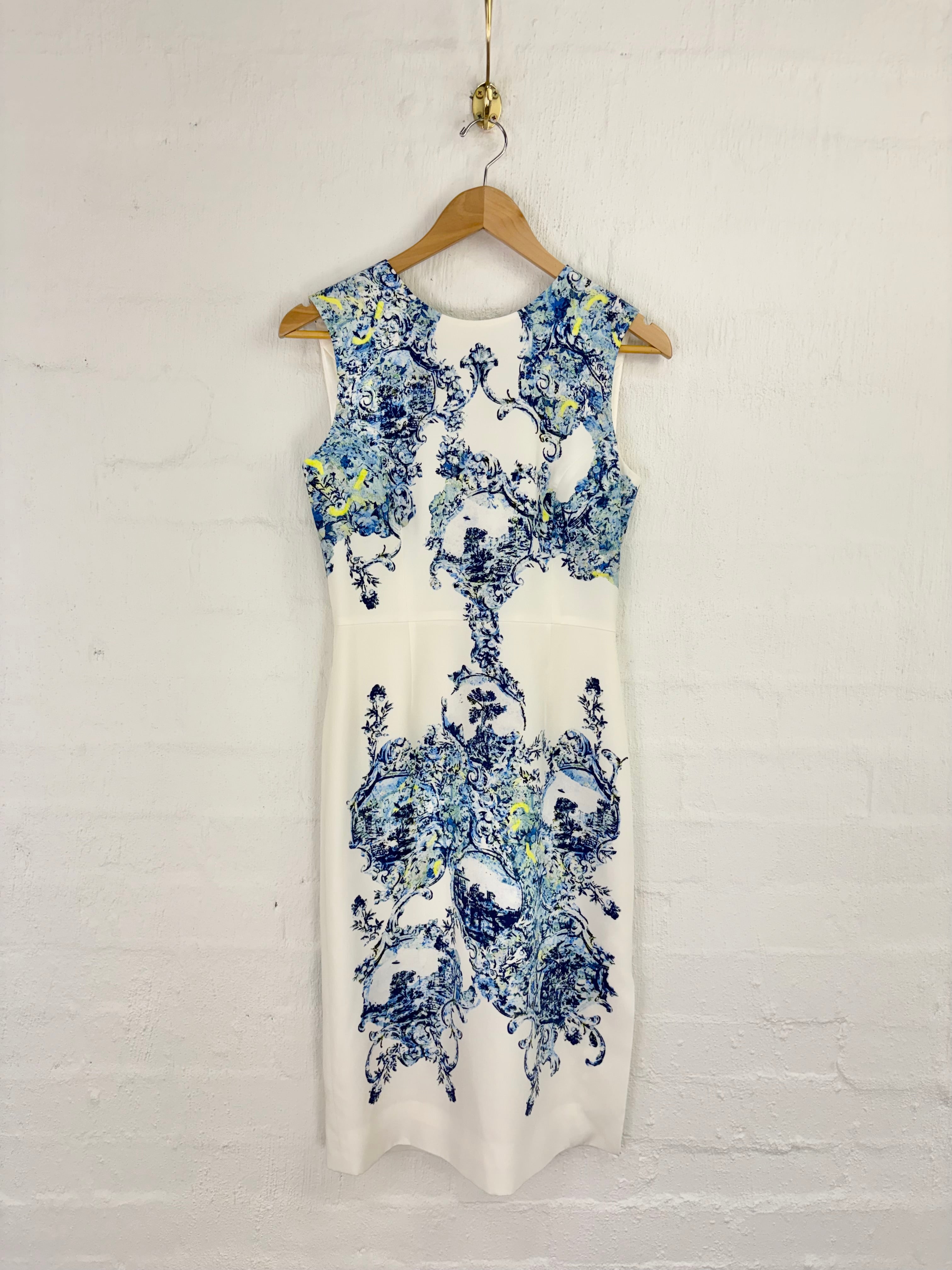 A sleeveless white dress with a blue and yellow floral toile pattern, hanging on a wooden hanger against a white painted brick wall.