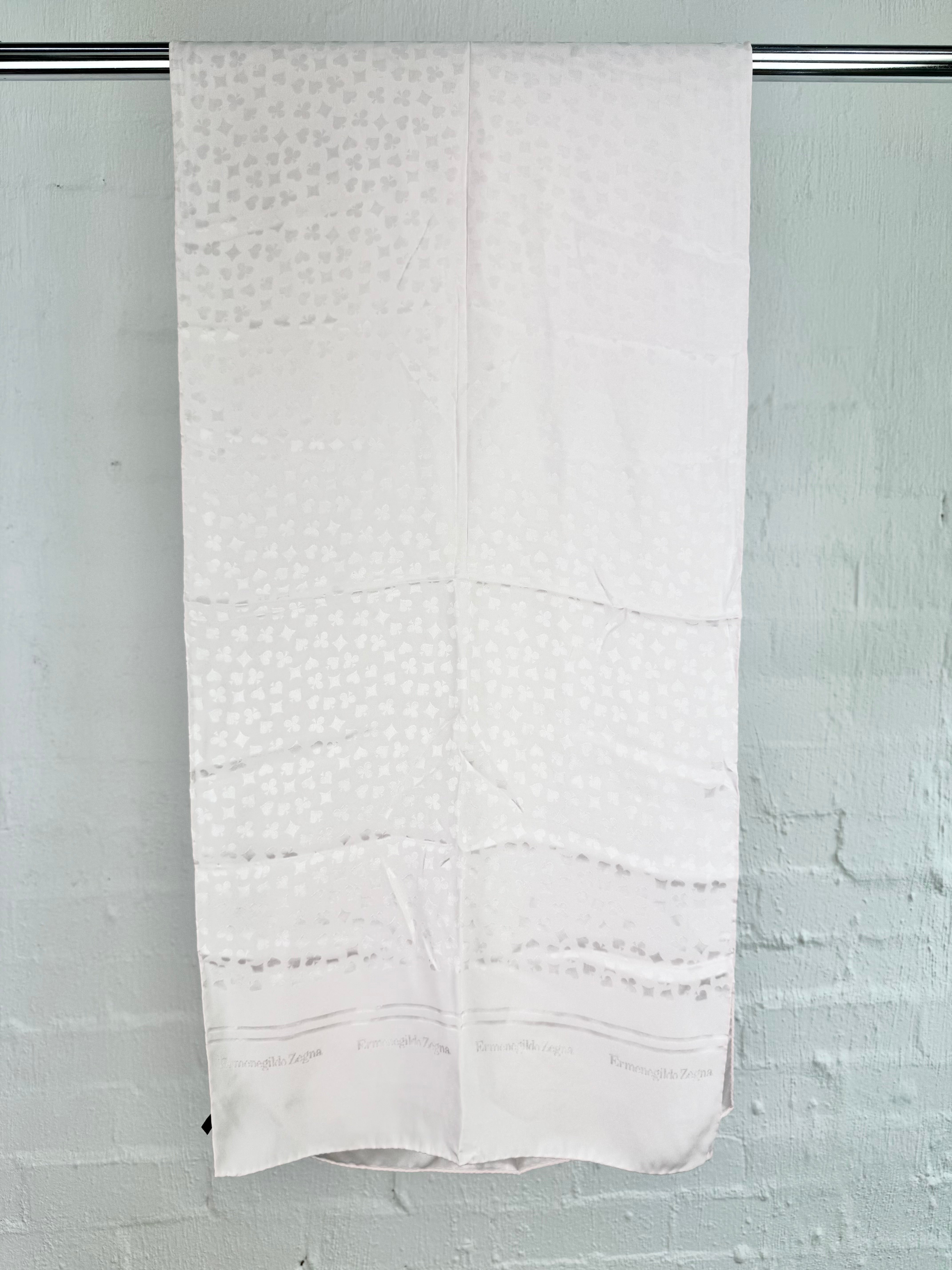A white patterned fabric panel or curtain hanging from a metal rod against a white painted brick wall.