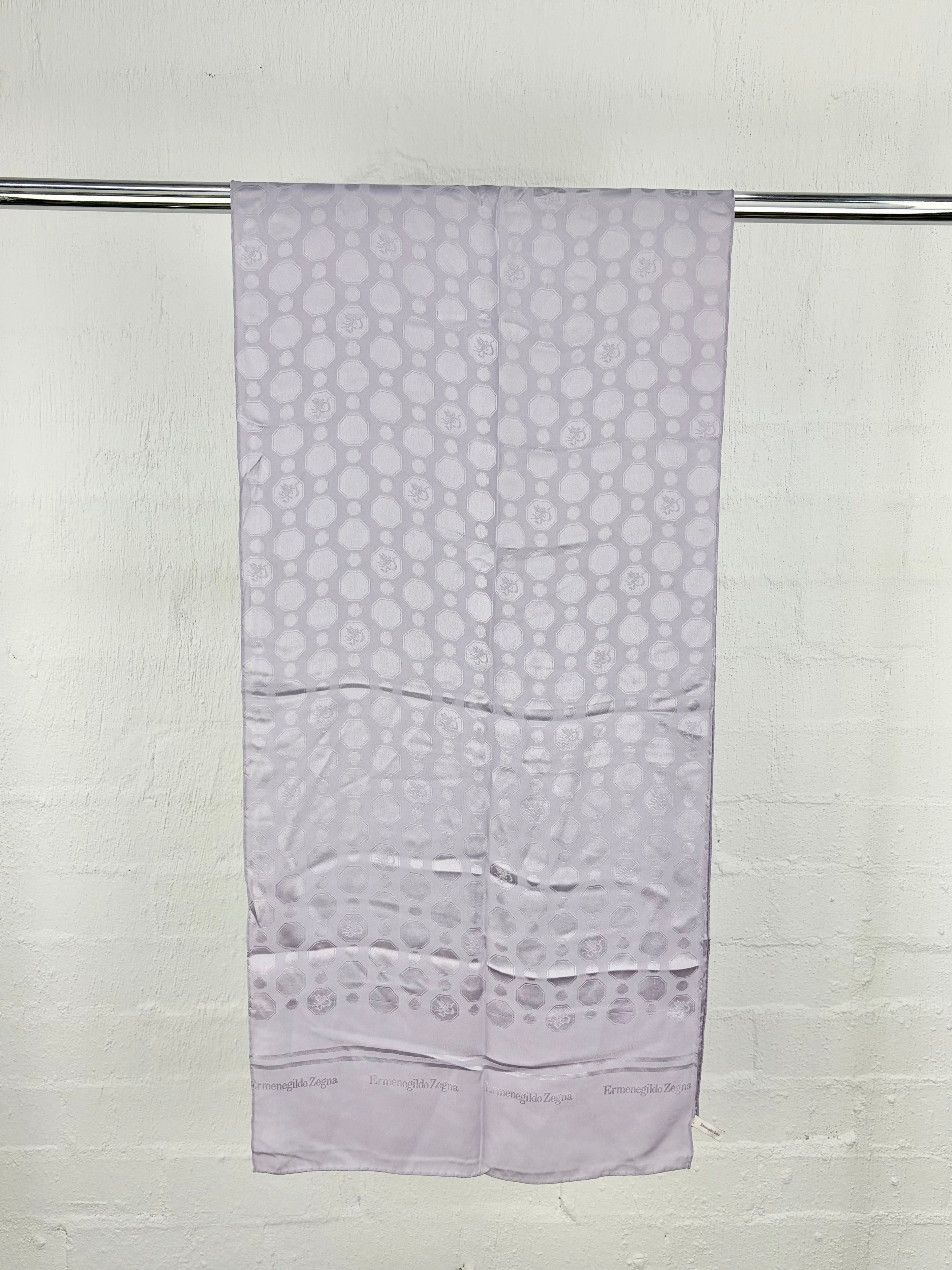 A light lavender scarf with a repeating circular pattern and a decorative border, hanging from a metal rod against a white brick wall.