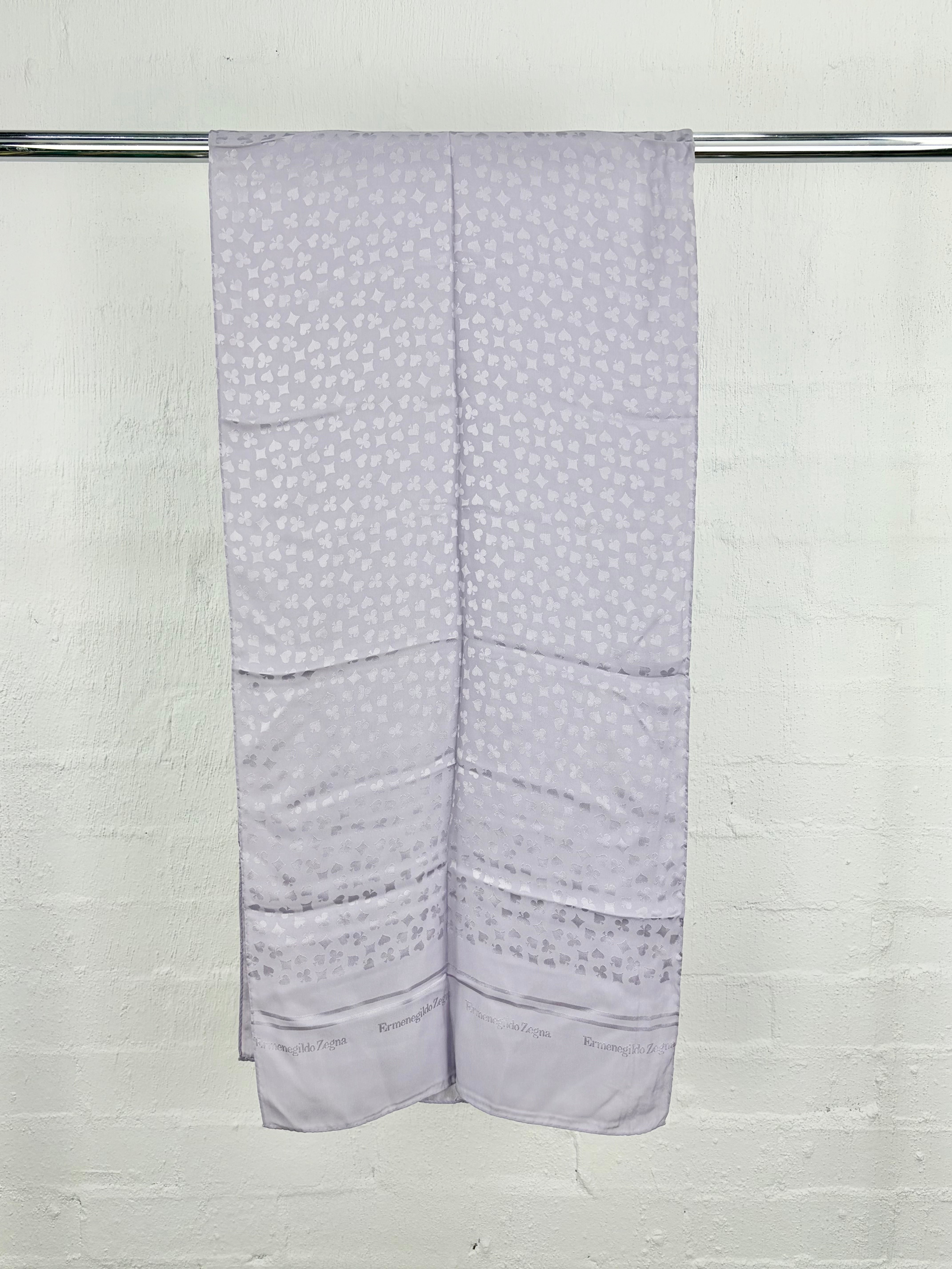 A light lavender-gray patterned scarf draped over a metal rod against a white painted brick wall.