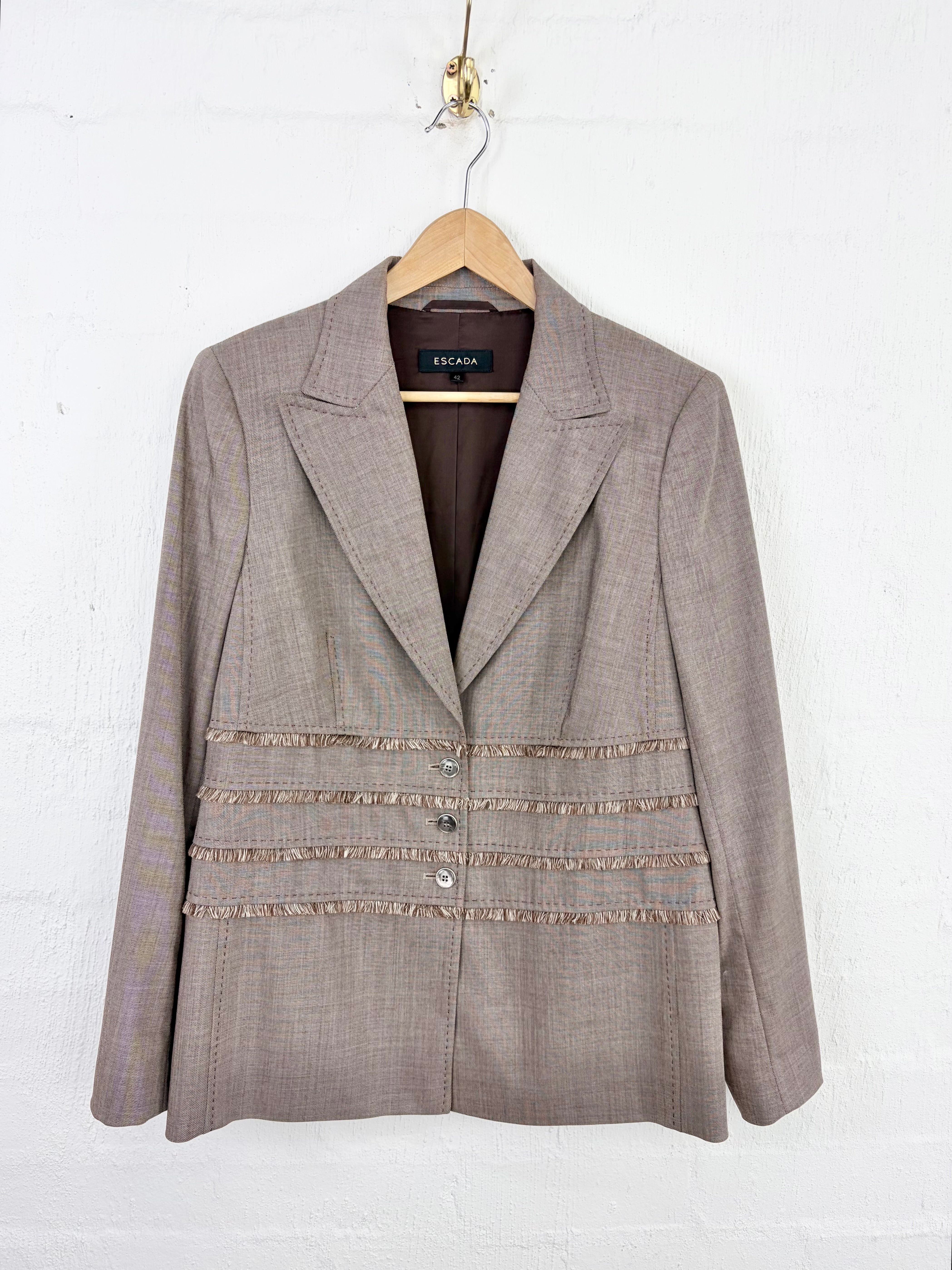 A taupe tweed blazer with decorative fringed horizontal stripes across the front, hanging on a wooden hanger against a white painted brick wall.
