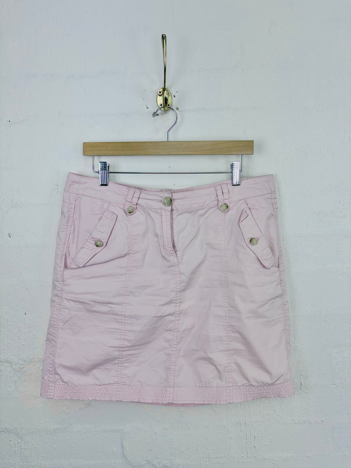 A light pink mini skirt with front button details and side pockets, hanging from a wooden hanger on a white textured wall.