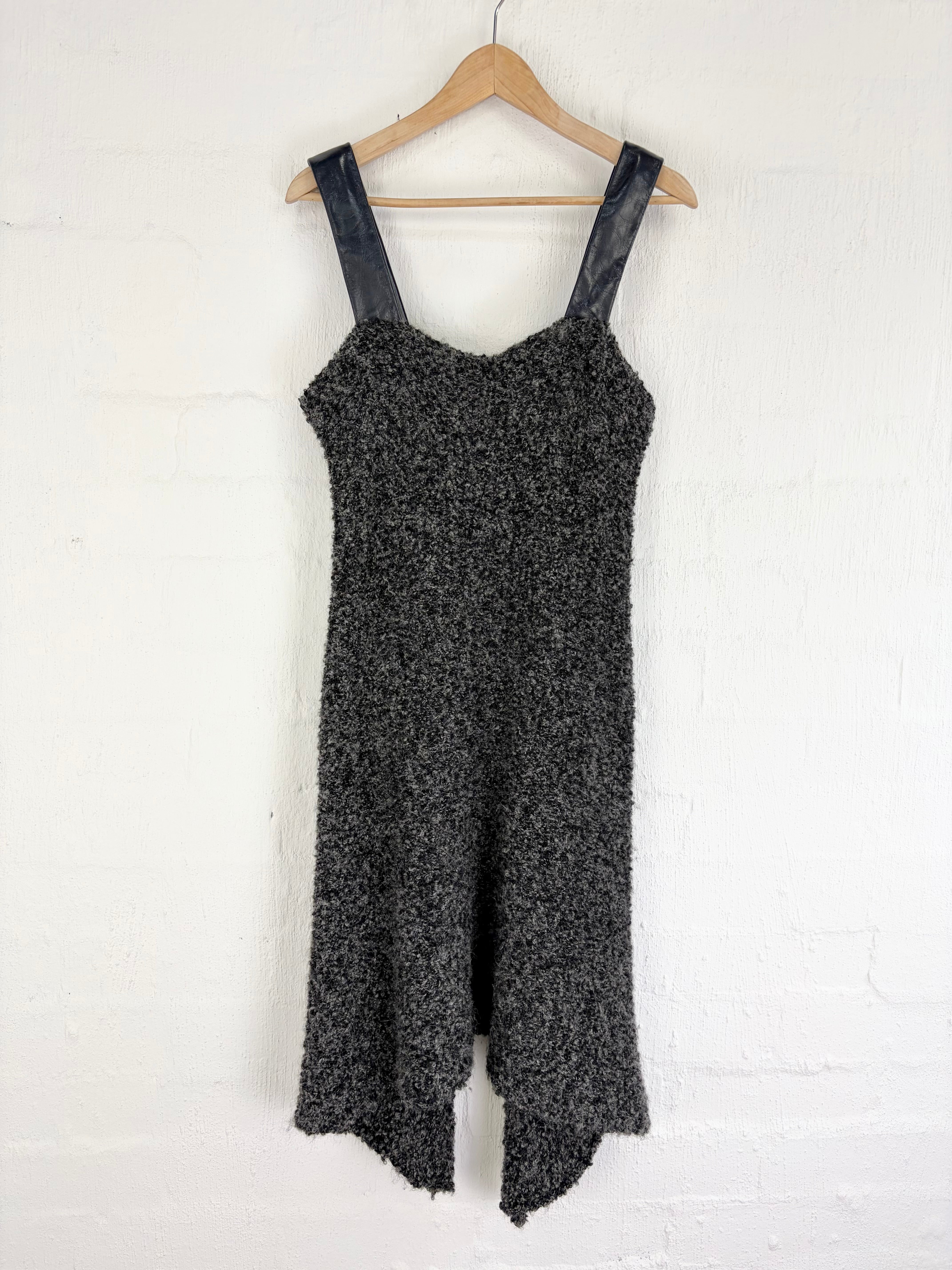 A sleeveless black and gray marled knit jumpsuit with dark faux-leather shoulder straps, hanging on a wooden hanger against a white brick wall.