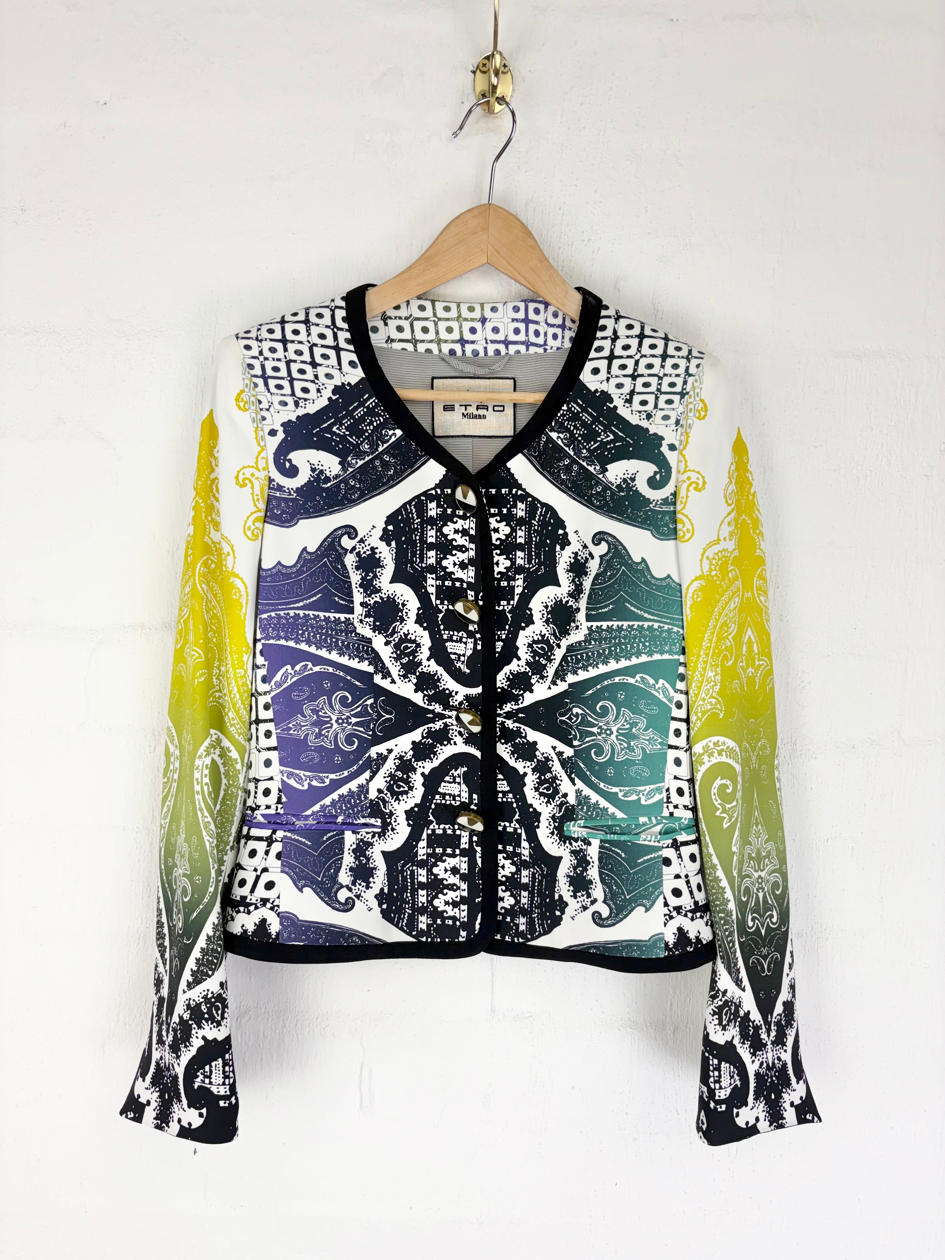 A cropped long-sleeve jacket with a complex multicolor pattern featuring black, white, purple, teal, and yellow gradients, hanging on a wooden hanger against a white brick wall.