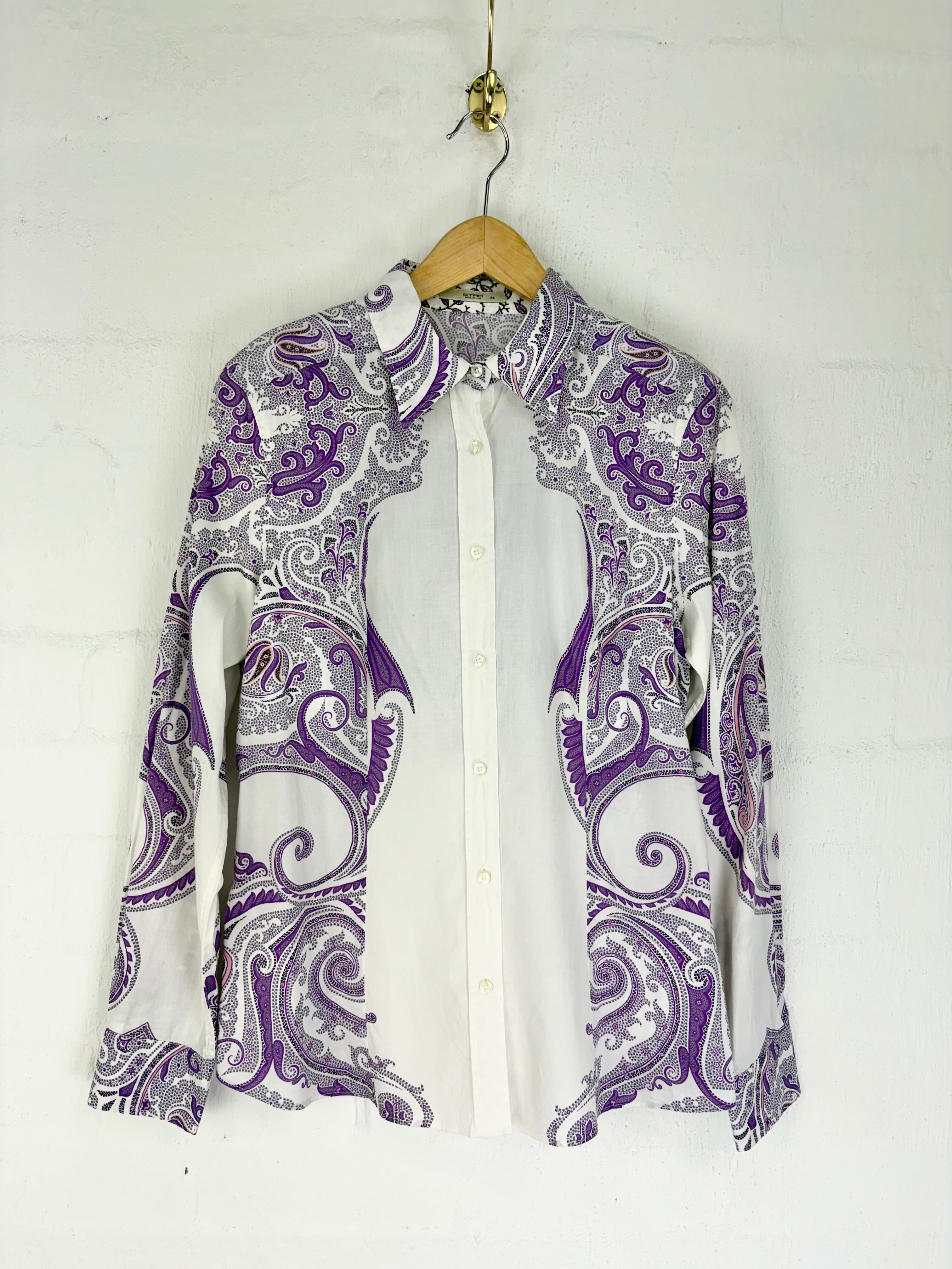 A white button-down shirt with a purple and gray paisley pattern, hanging on a wooden hanger against a white brick wall.