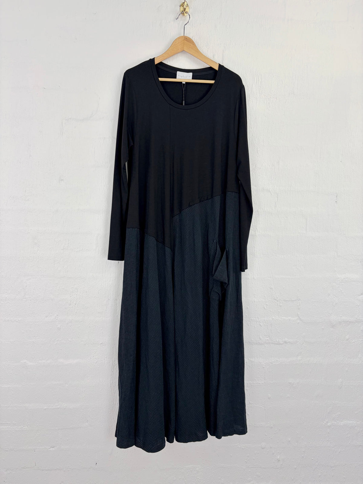 A long-sleeve midi dress in black and dark navy blue, hanging on a wooden hanger against a white painted brick wall.