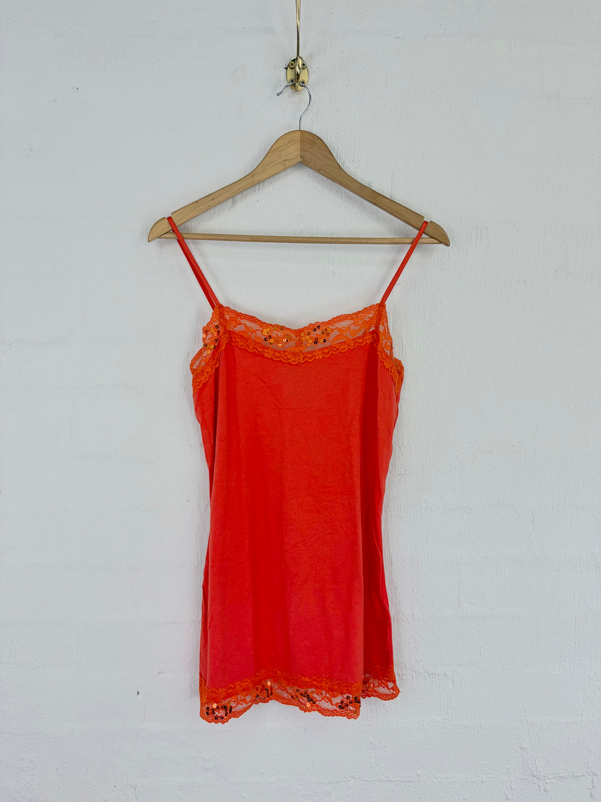 A bright red-orange sheer camisole slip with floral lace trim at the neckline and hem, hanging on a wooden hanger against a white painted brick wall.