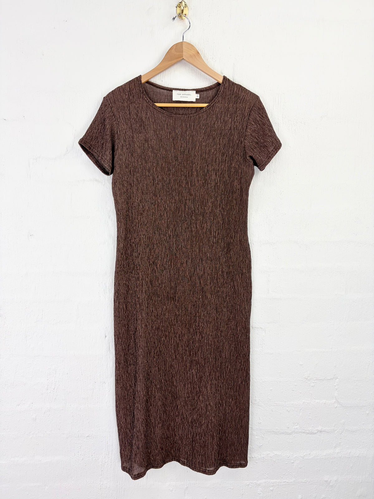 A short-sleeve brown knit dress with a textured, heathered finish, hanging on a wooden hanger against a white painted brick wall.