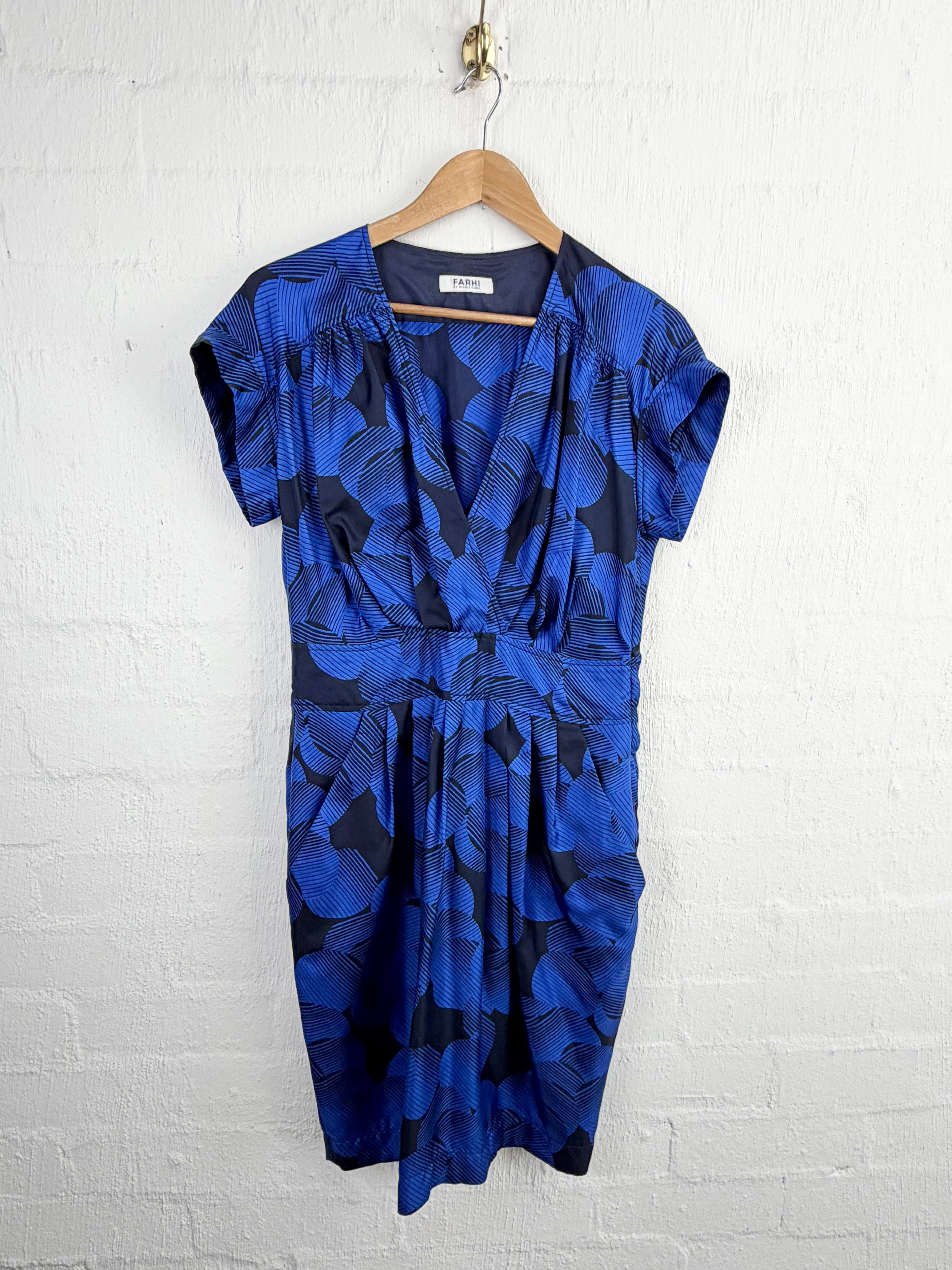 A blue and black abstract patterned wrap dress with short sleeves, hanging on a wooden hanger against a white painted brick wall.