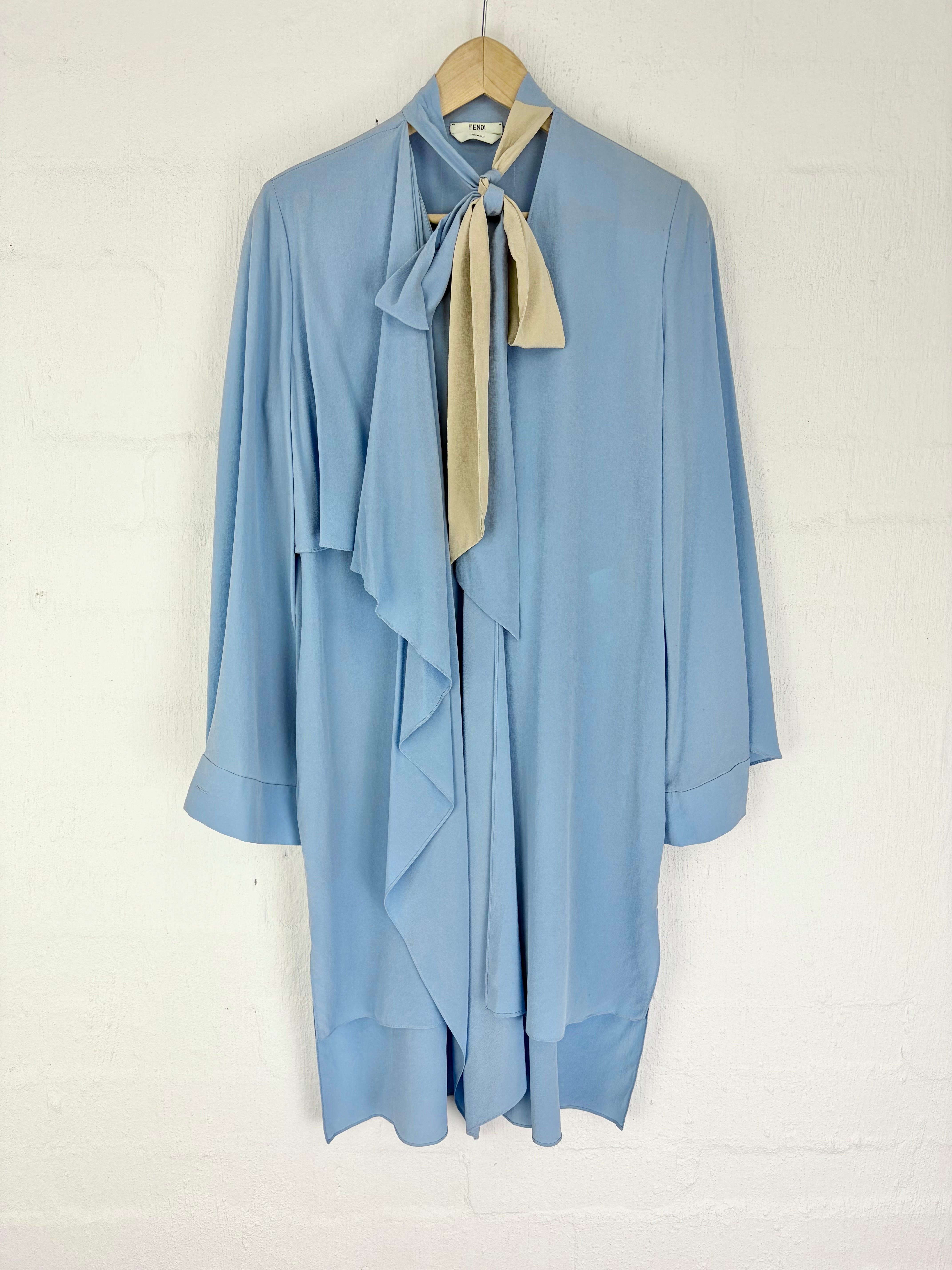 A light blue long-sleeve blouse with a draped, layered ruffle design and a beige satin necktie bow at the collar, hanging on a wooden hanger against a white painted brick wall.