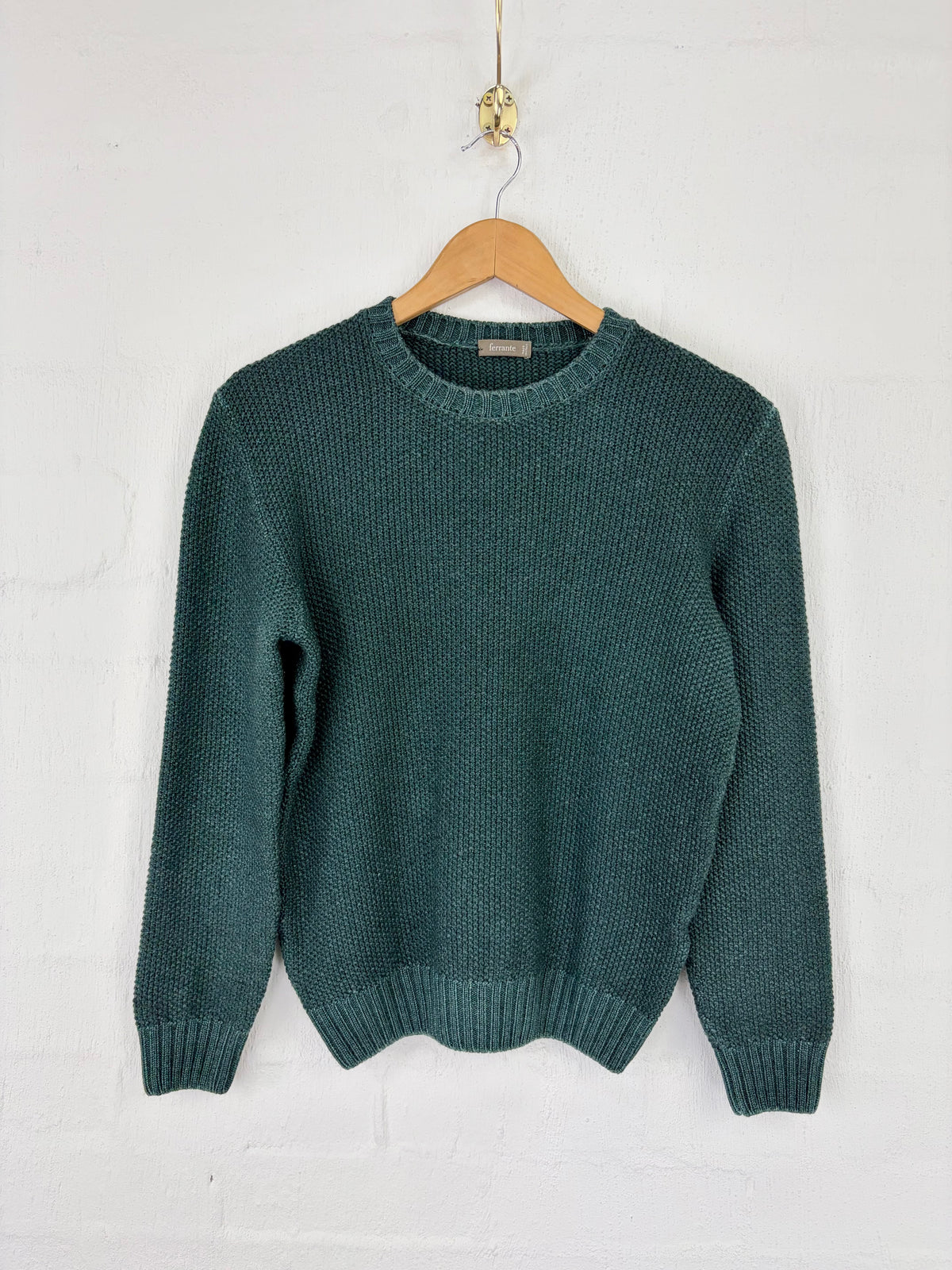 A dark teal green knit crewneck sweater hanging on a wooden hanger against a white painted brick wall.