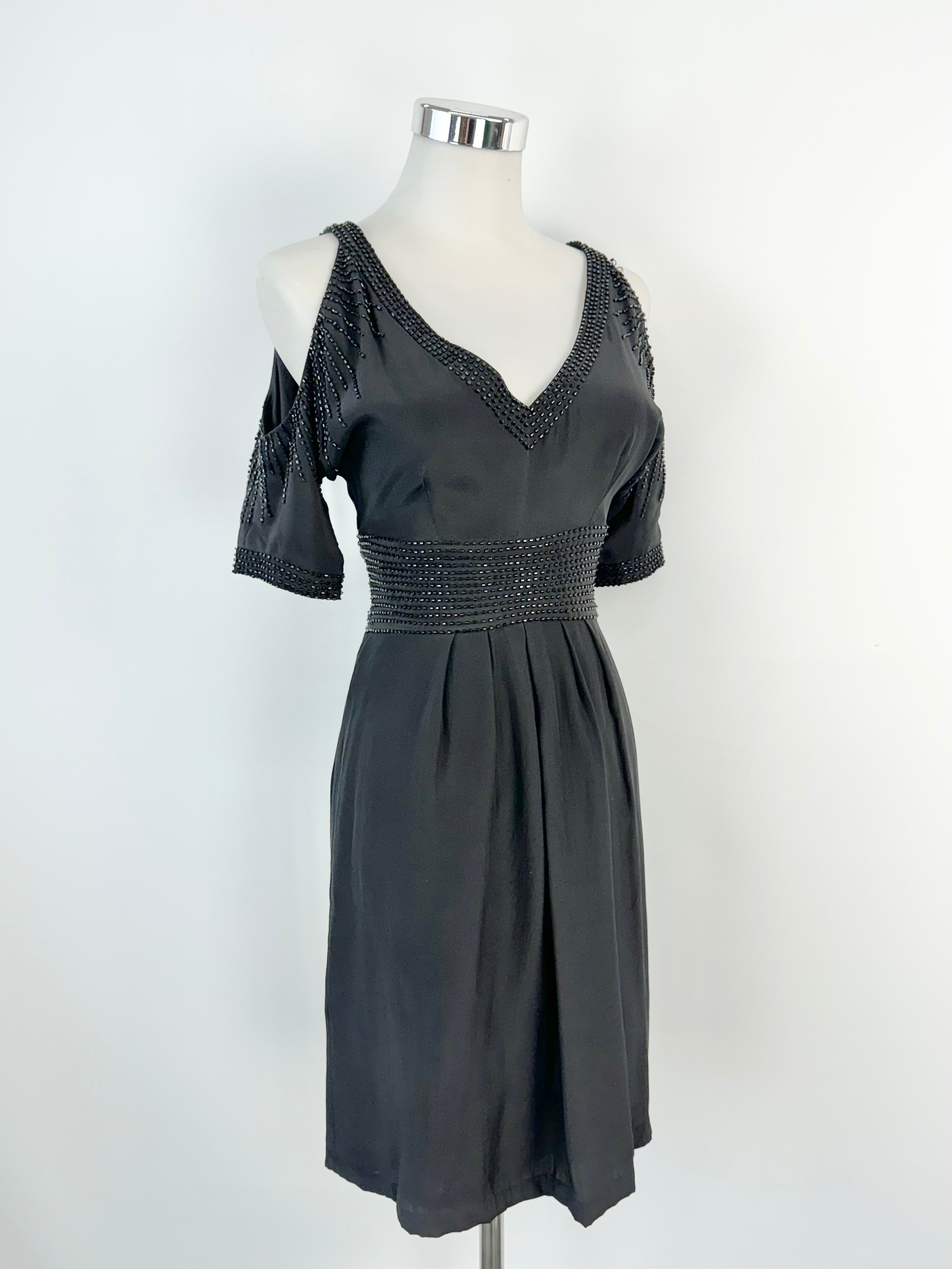 A black beaded midi dress with cut out shoulders and a v-neckline, displayed on a mannequin against a white background.