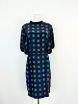 A silk sheath dress with a retro geometric pattern in blue and orange, featuring small buttons on the waist, a high neckline, and a wide rib knit trim.