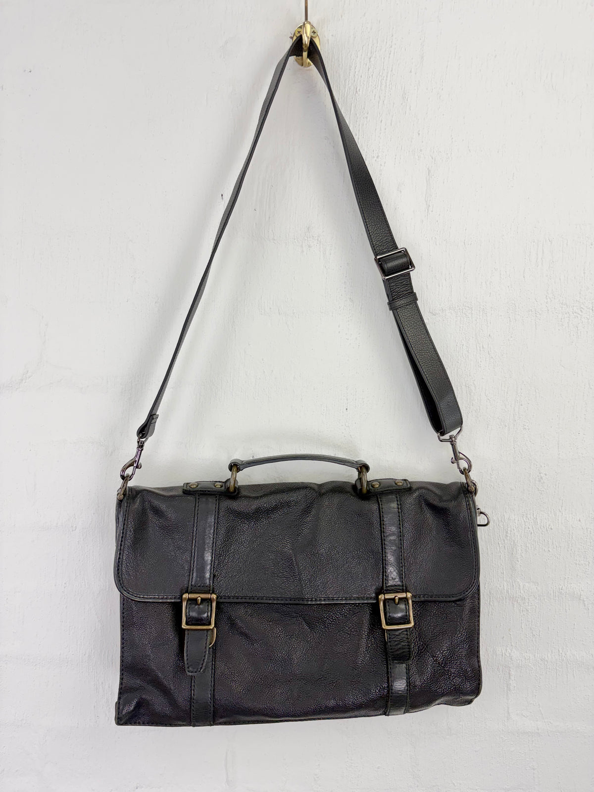 A black leather briefcase-style shoulder bag with a top handle, adjustable shoulder strap, and two buckle details on the front flap, hanging against a white wall.