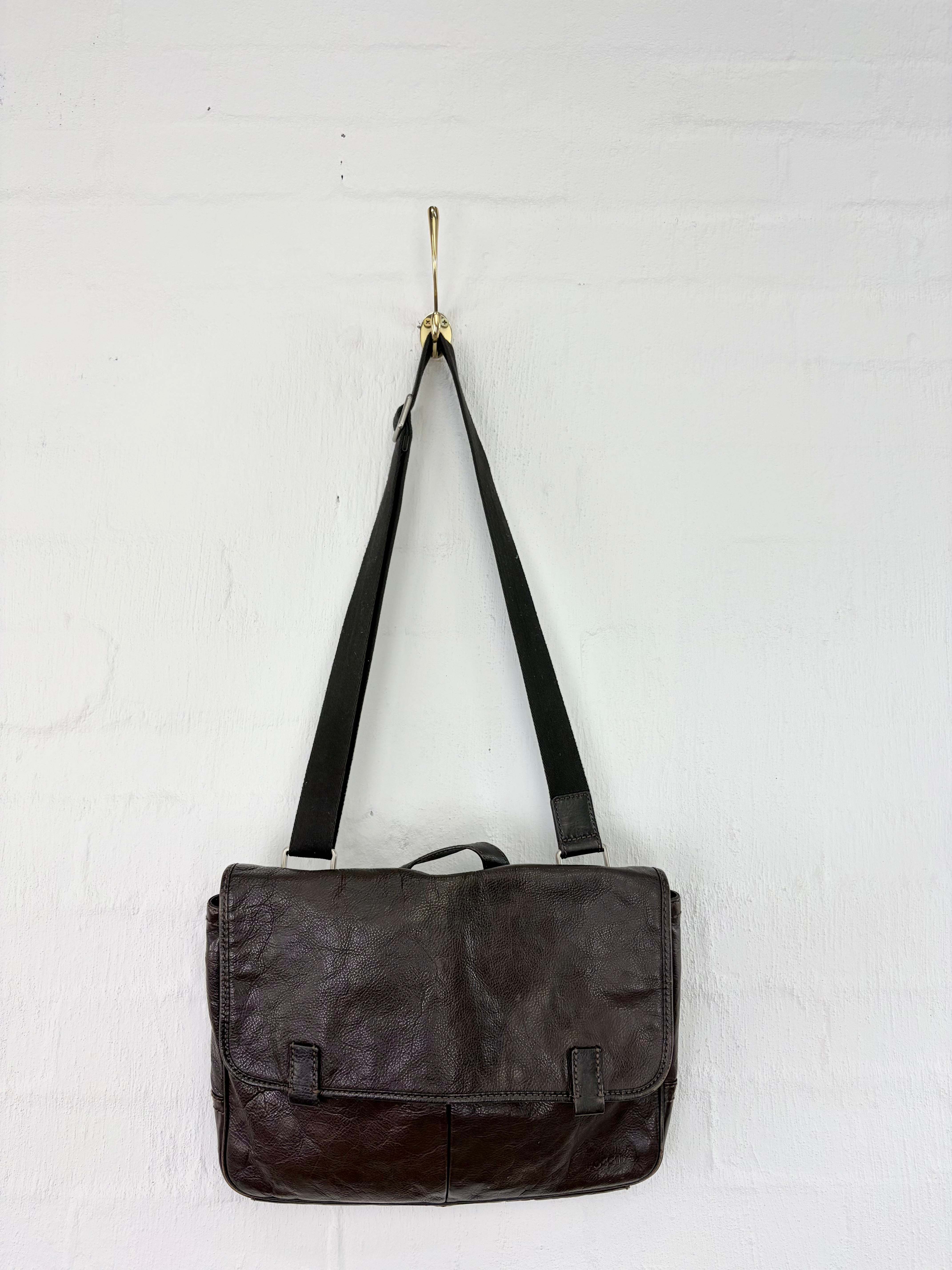 A dark brown leather messenger bag with a black shoulder strap, hanging from a gold hook on a white painted brick wall.