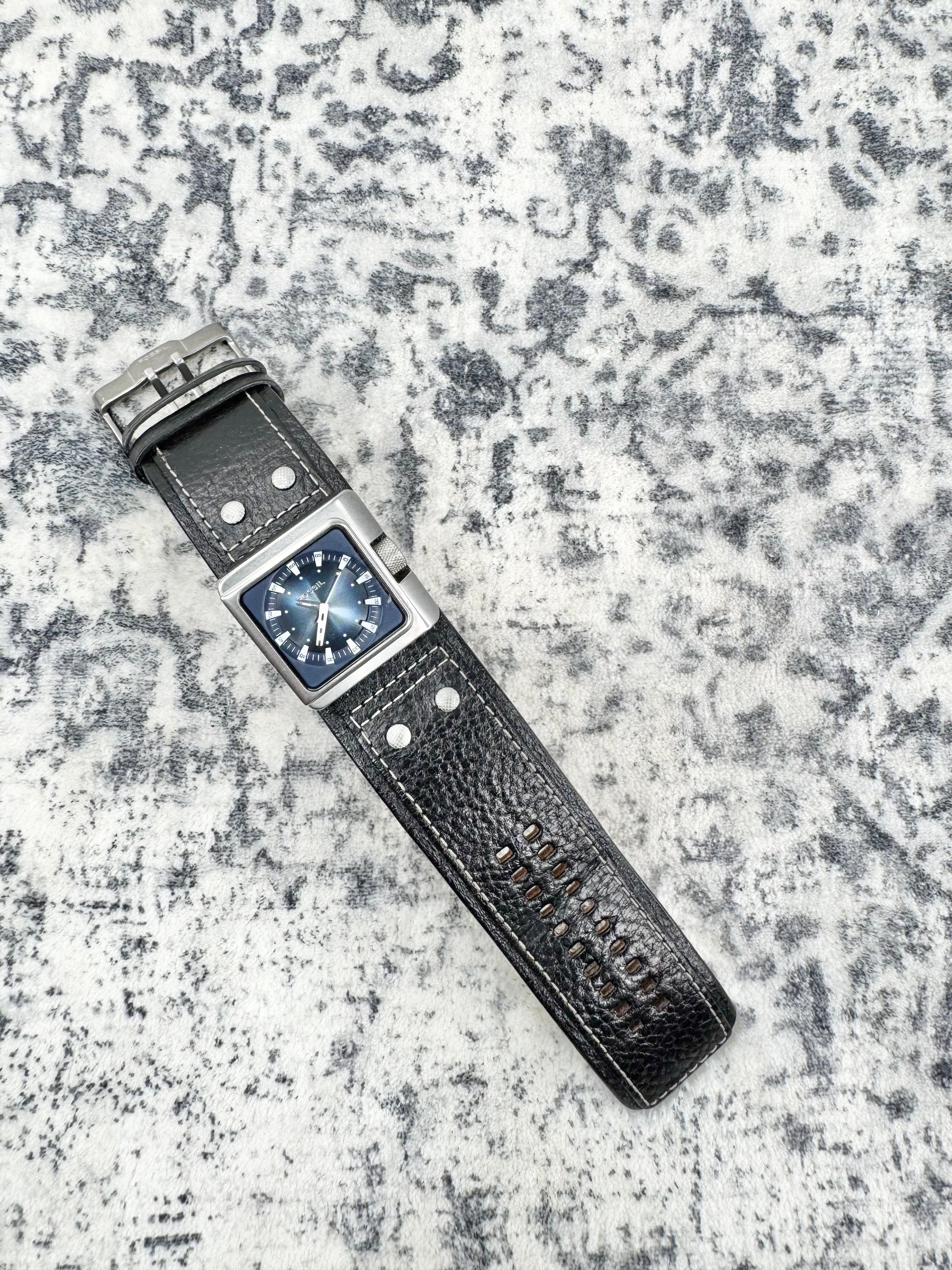 A rectangular silver-tone metal watch with a dark blue dial and a textured black leather strap, laid diagonally on a mottled gray and white textured surface.