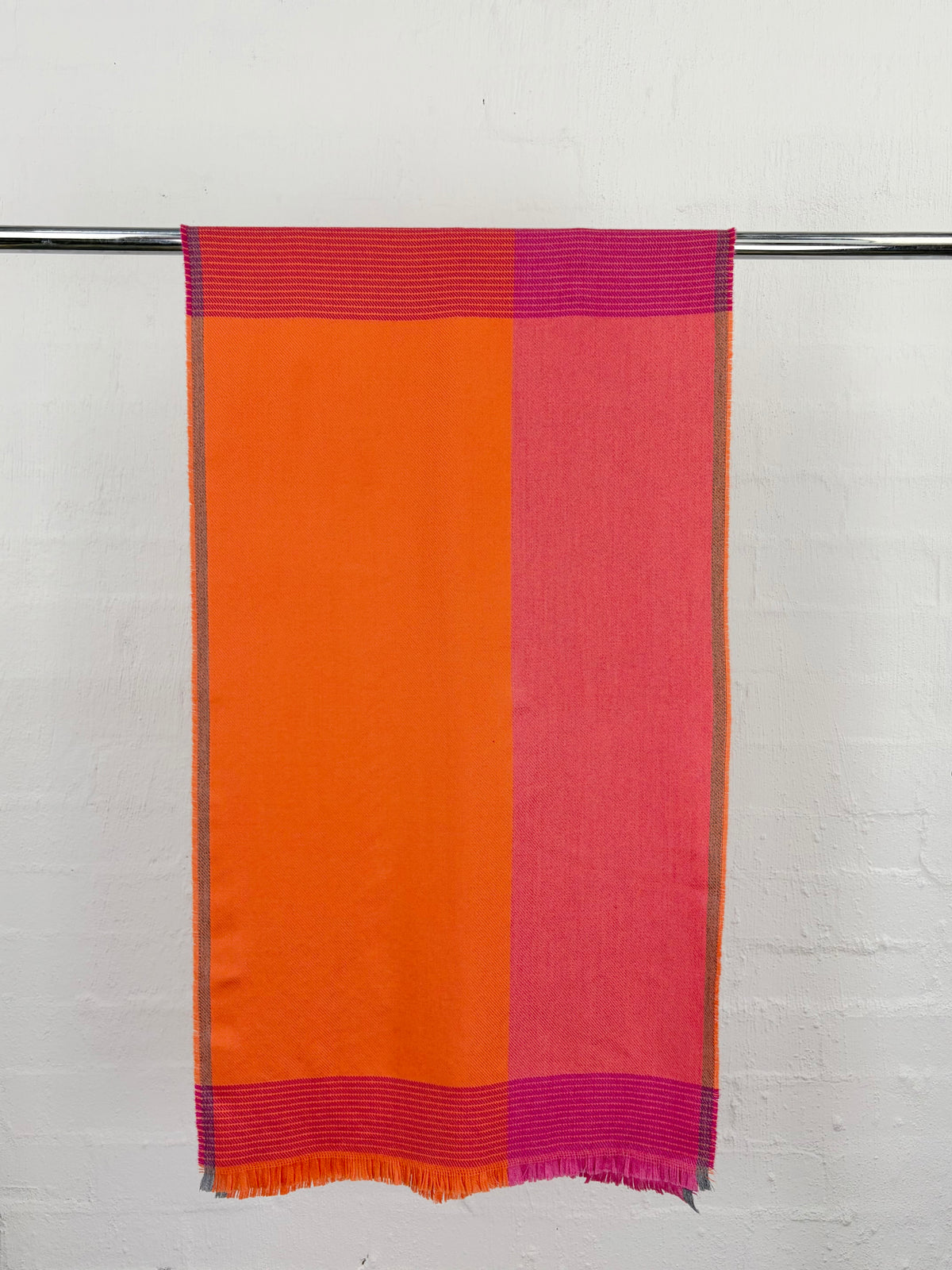 A rectangular woven textile, likely a scarf or throw, hanging from a metal rod against a white painted brick wall. The fabric features a bold color-blocked design with a large orange panel on the left, a pink panel on the right, and magenta striped borders at the top and bottom.