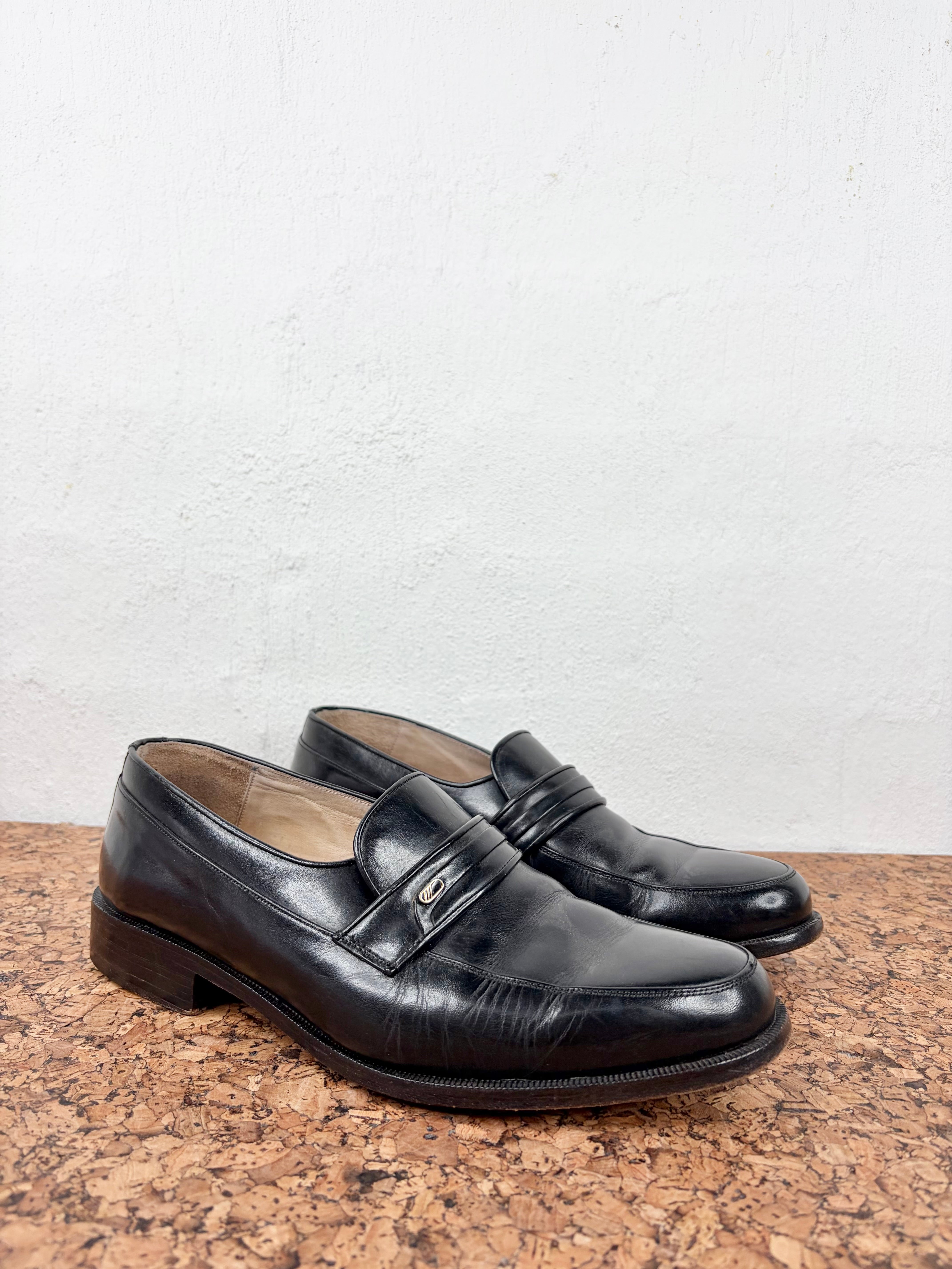 A pair of black leather penny loafers with a glossy finish, resting on a brown cork surface against a white textured wall.