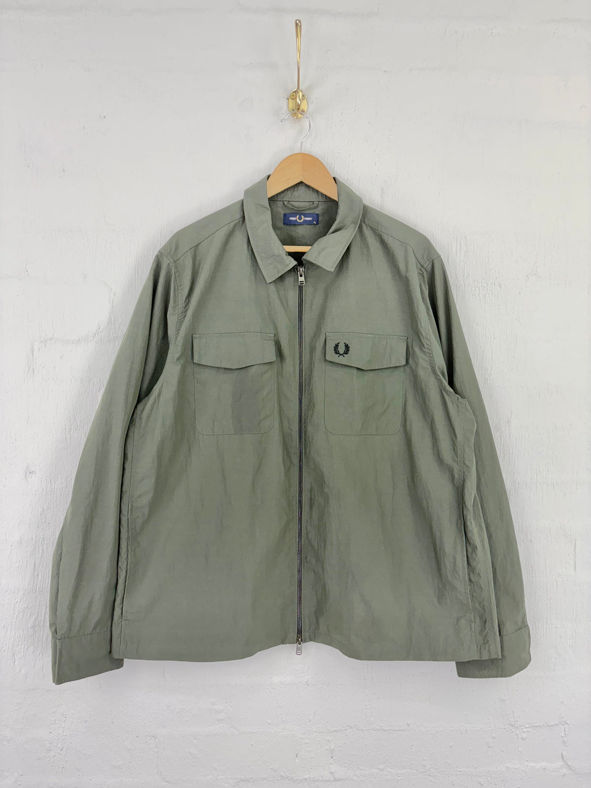 An olive green zip-up jacket by Fred Perry hangs on a wooden hanger against a white painted brick wall. The jacket features a classic collar, a front zipper, and two flap chest pockets, with the iconic embroidered laurel wreath logo on the left chest.
