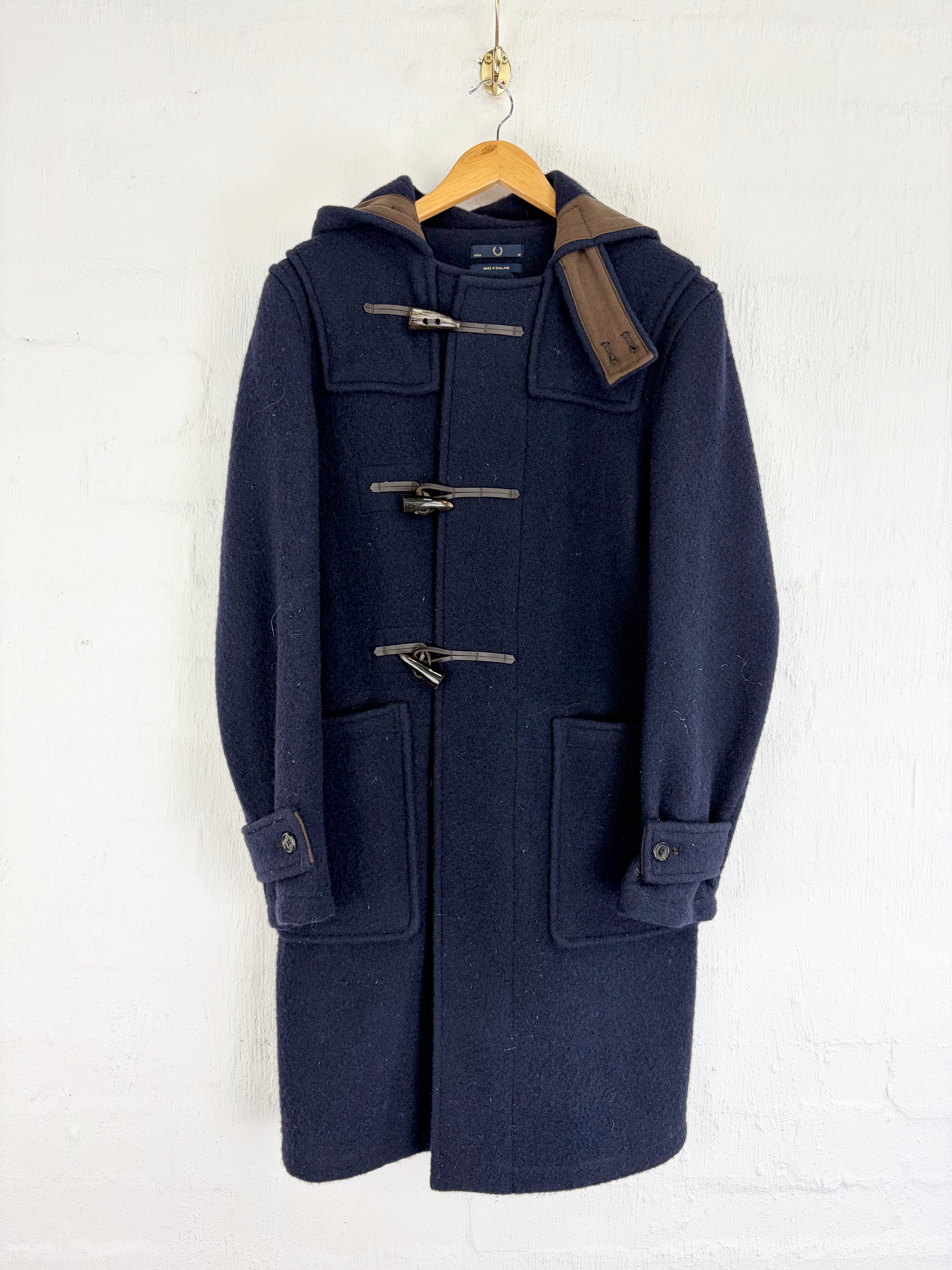 A navy blue wool duffle coat with brown corduroy hood lining and toggle closures, hanging on a wooden hanger against a white painted brick wall.