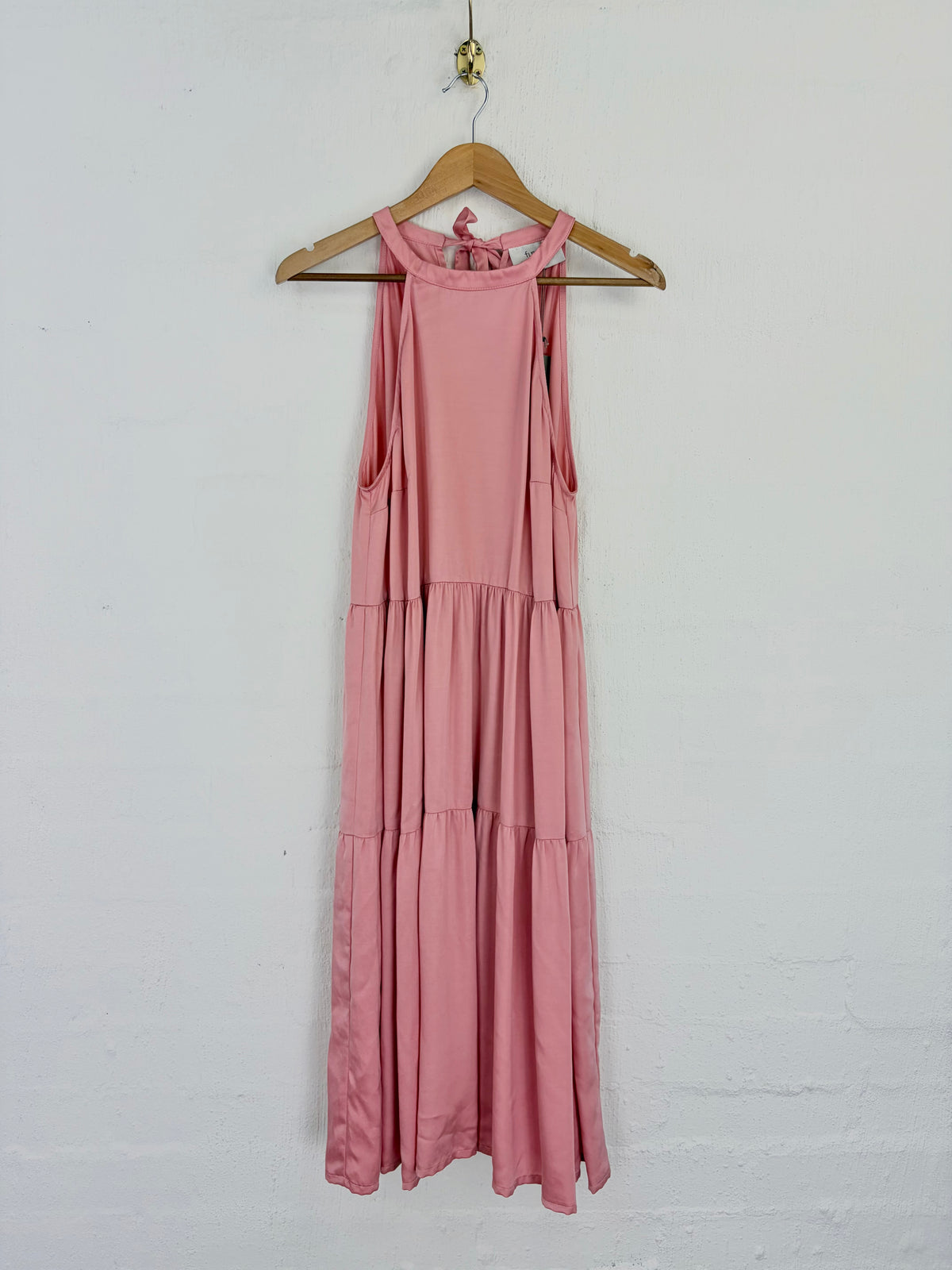 A sleeveless tiered maxi dress in dusty pink, hanging on a wooden hanger against a white painted brick wall.