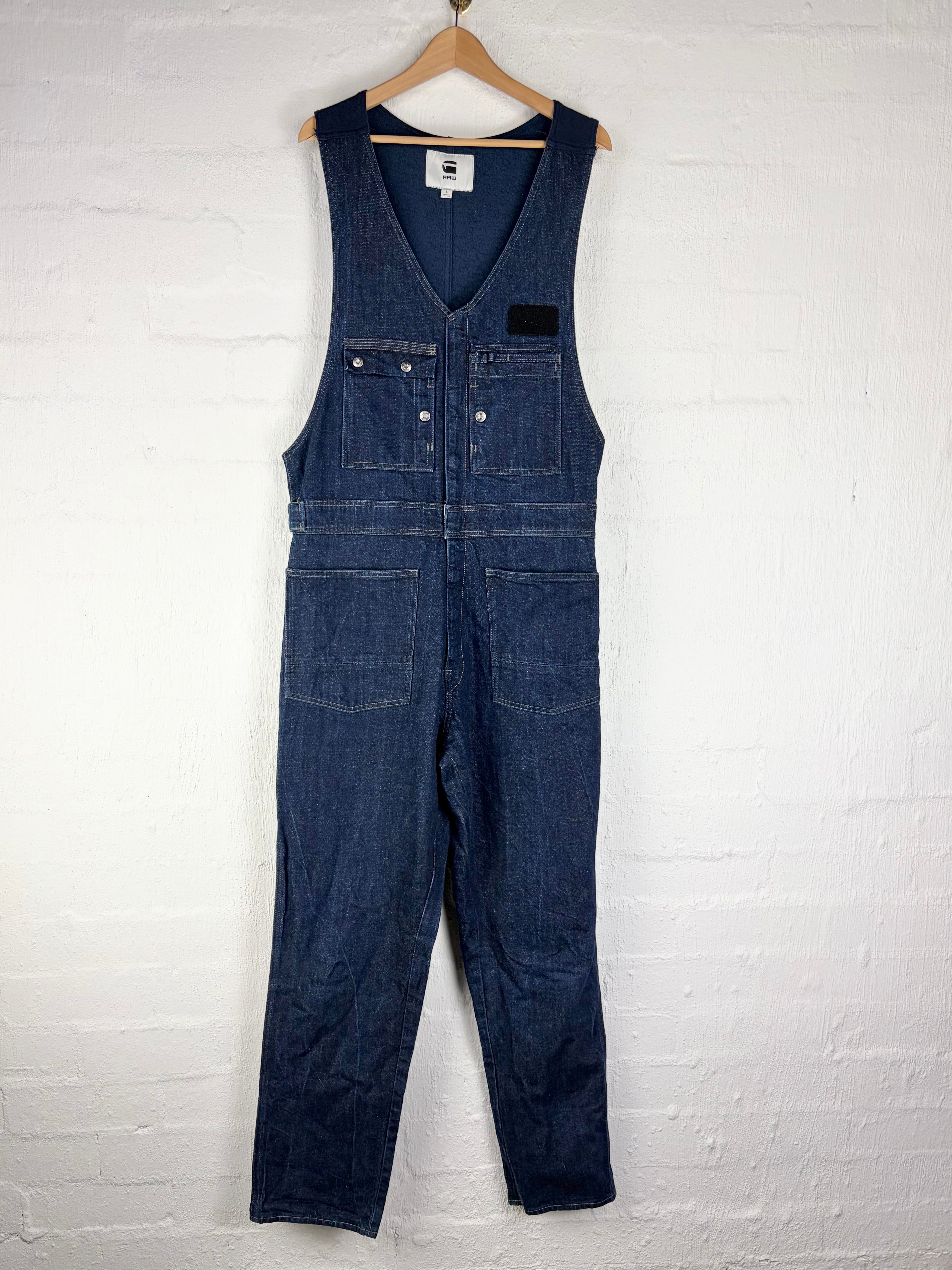 A pair of dark blue denim bib overalls hanging on a wooden hanger against a white painted brick wall.