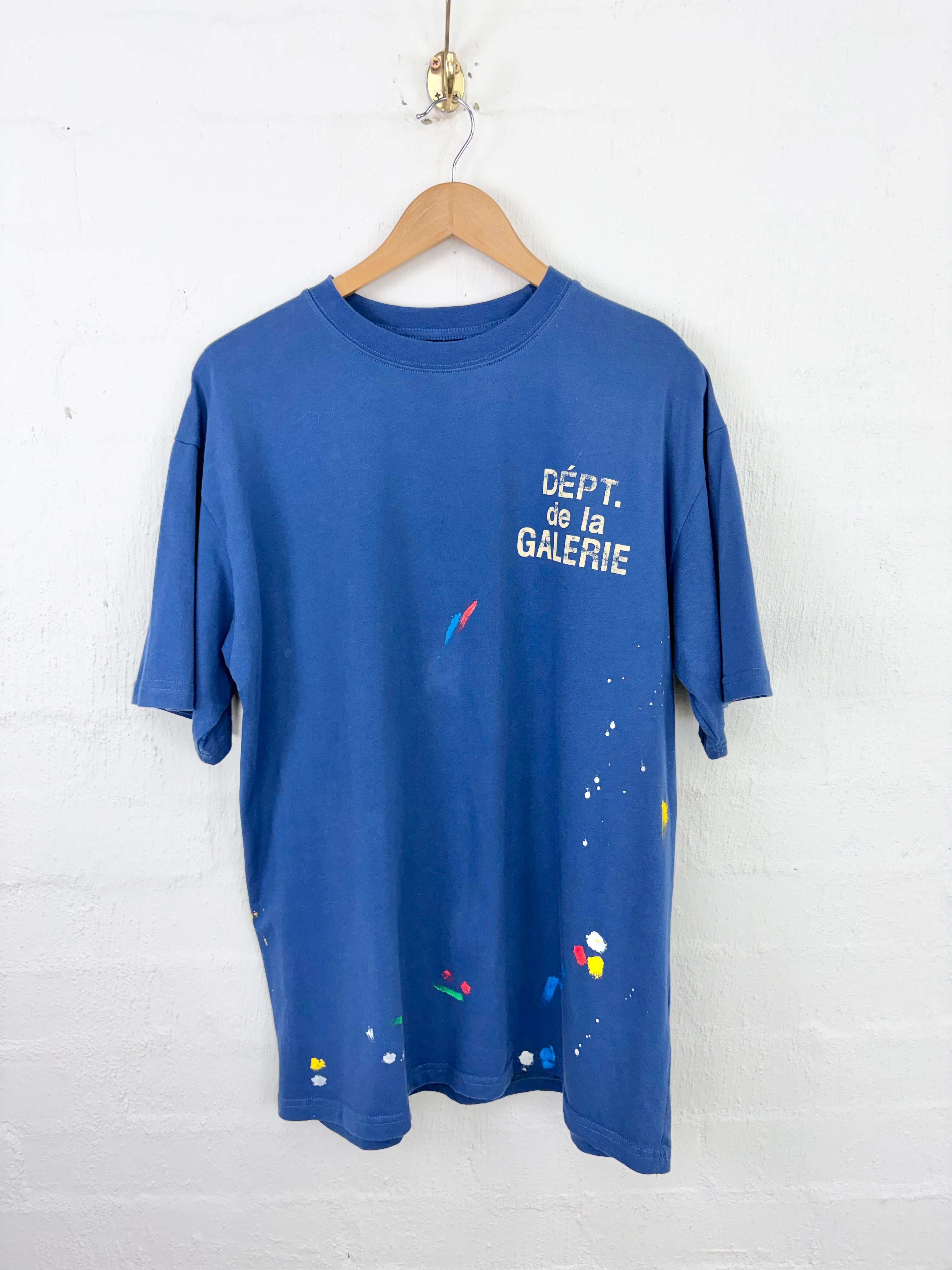 A blue short-sleeve t-shirt hanging on a wooden hanger against a white painted brick wall. The shirt features white text reading 'DÉPT.