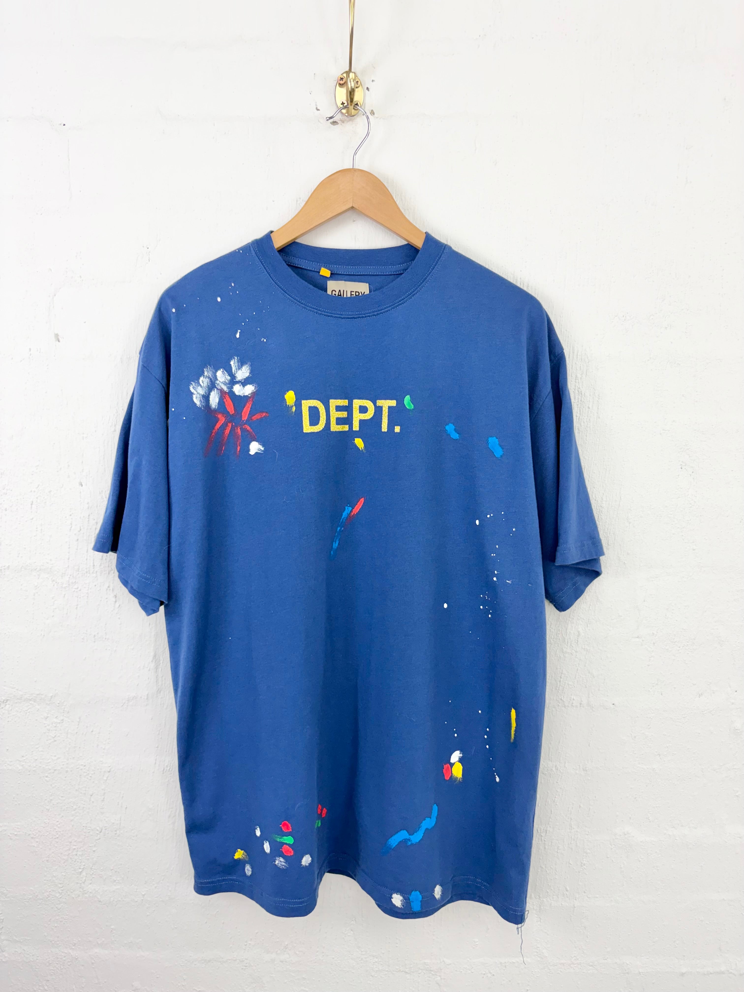 A blue short-sleeve t-shirt with a colorful paint splatter design and the text 'DEPT.' printed in yellow on the chest, hanging on a wooden hanger against a white brick wall.