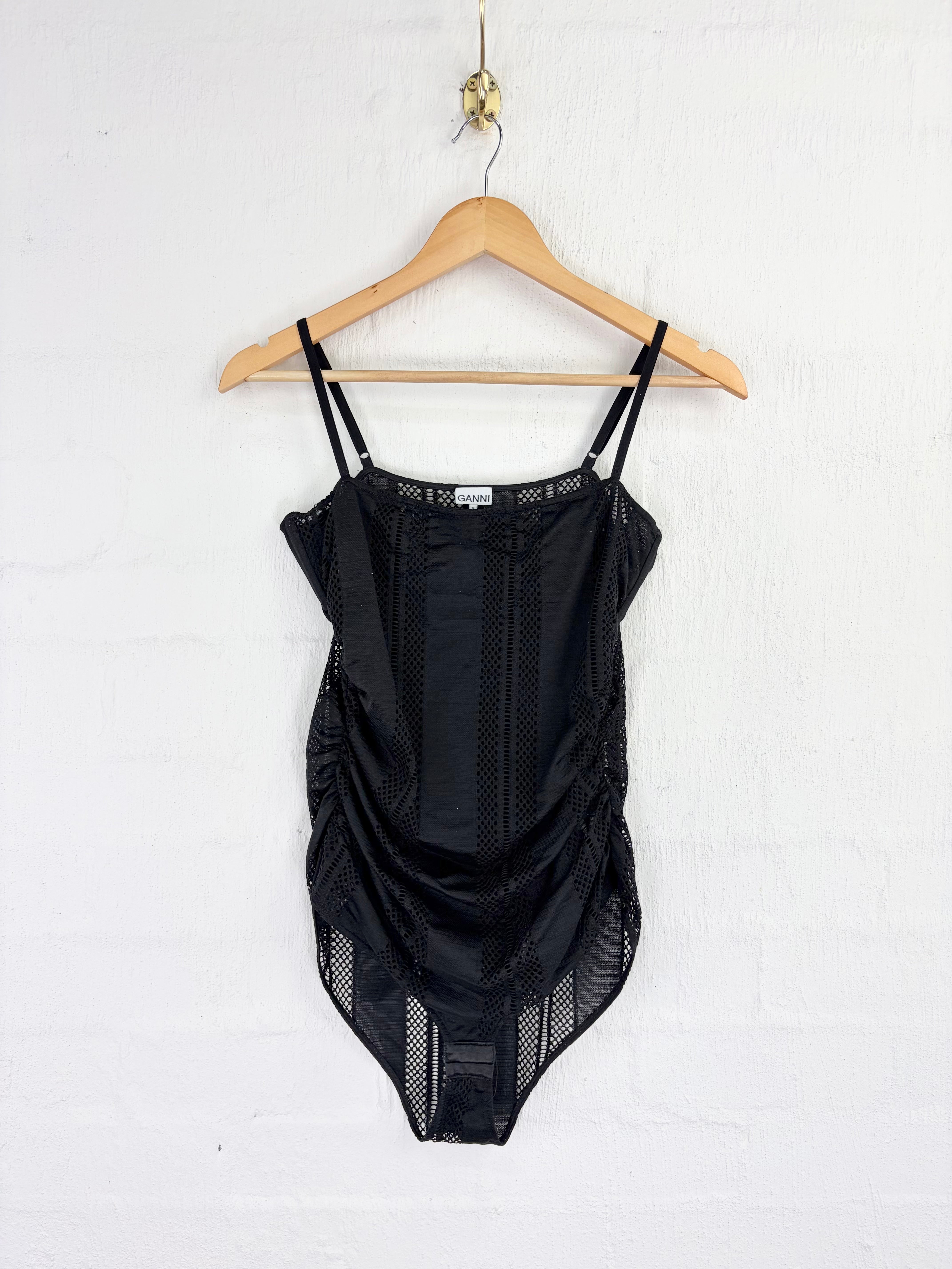 A black lace and mesh bodysuit with thin shoulder straps, hanging on a wooden hanger against a white painted brick wall.