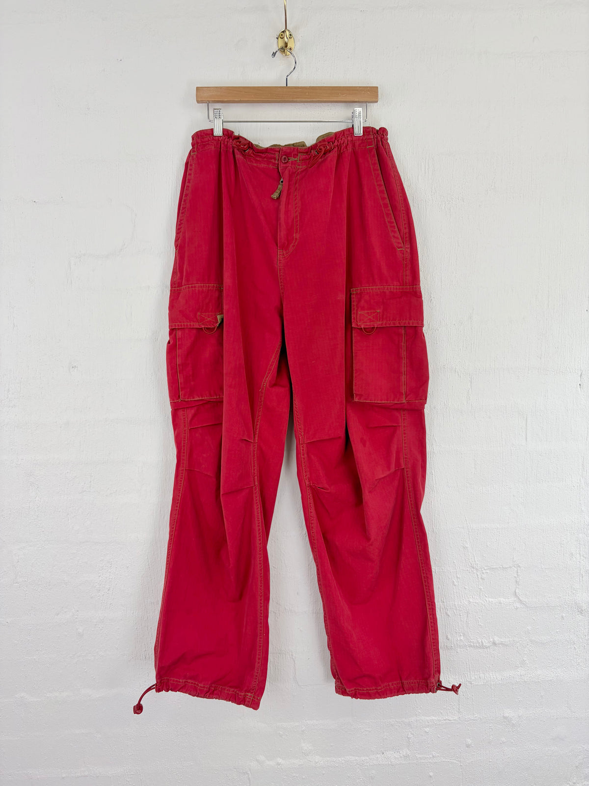 Red cargo pants with an elastic waistband and drawstring ankle cuffs, hanging from a wooden hanger against a white brick wall.