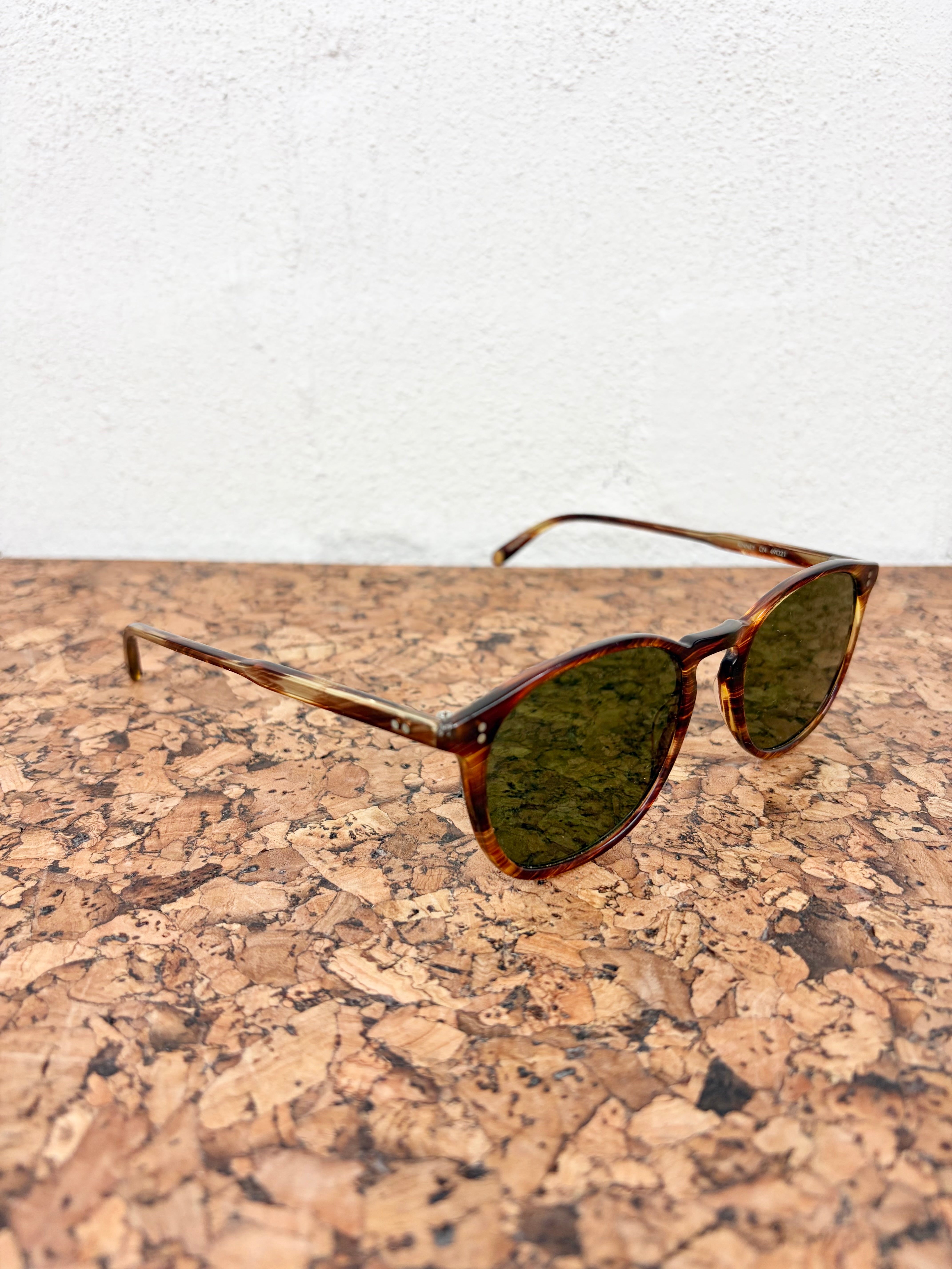 Oversized round sunglasses with tortoiseshell frames and dark green lenses, resting on a cork surface against a white wall.