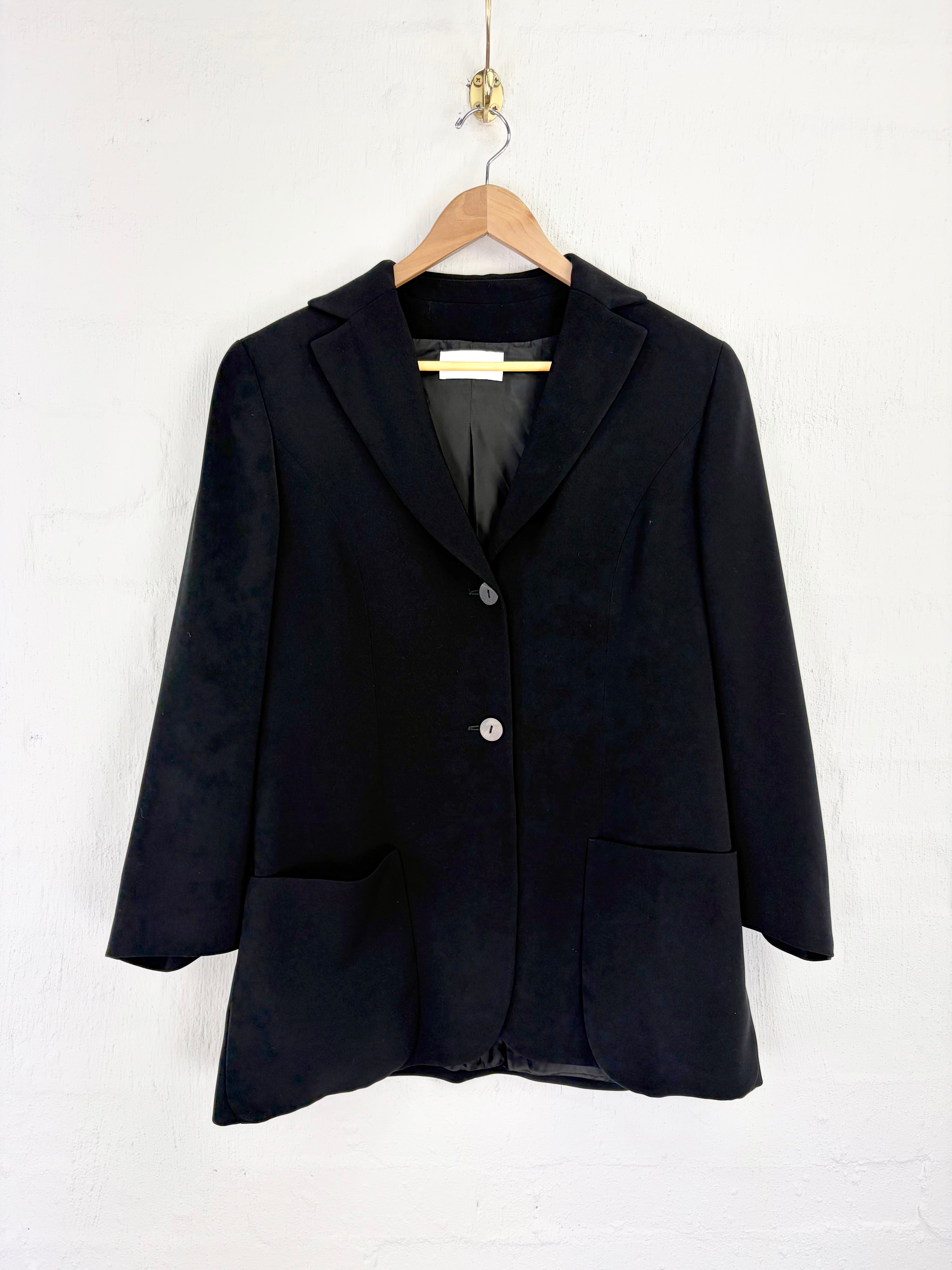 A black single-breasted blazer jacket with notch lapels and front patch pockets, hanging on a wooden hanger against a white textured wall.
