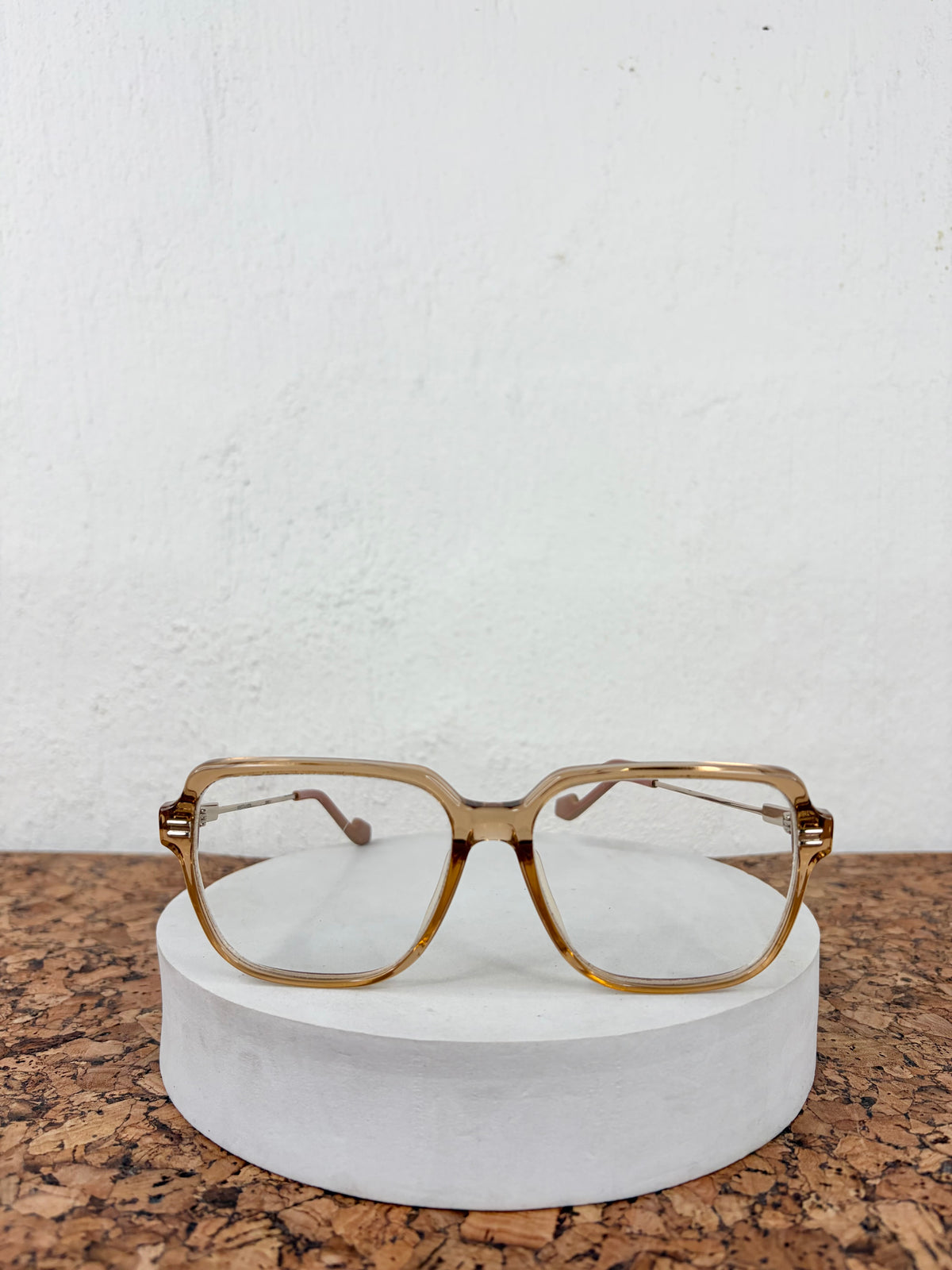 Vintage-style square eyeglasses with a translucent gold acetate frame, resting on a white cylindrical display stand against a white wall and cork surface.