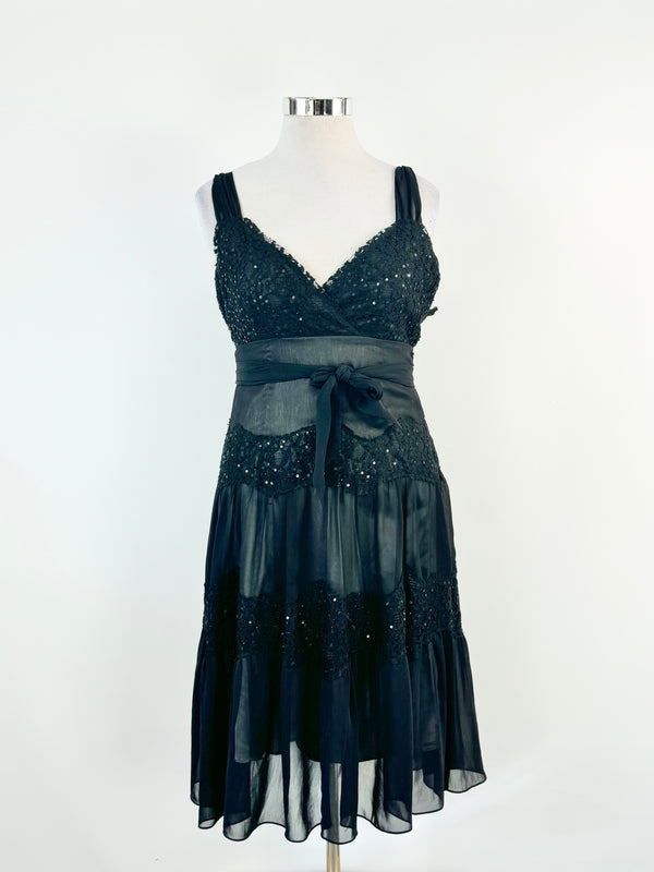 Black lace cocktail dress with sequins and metallic fabric, featuring adjustable straps and a tie at the waist, displayed on a mannequin.