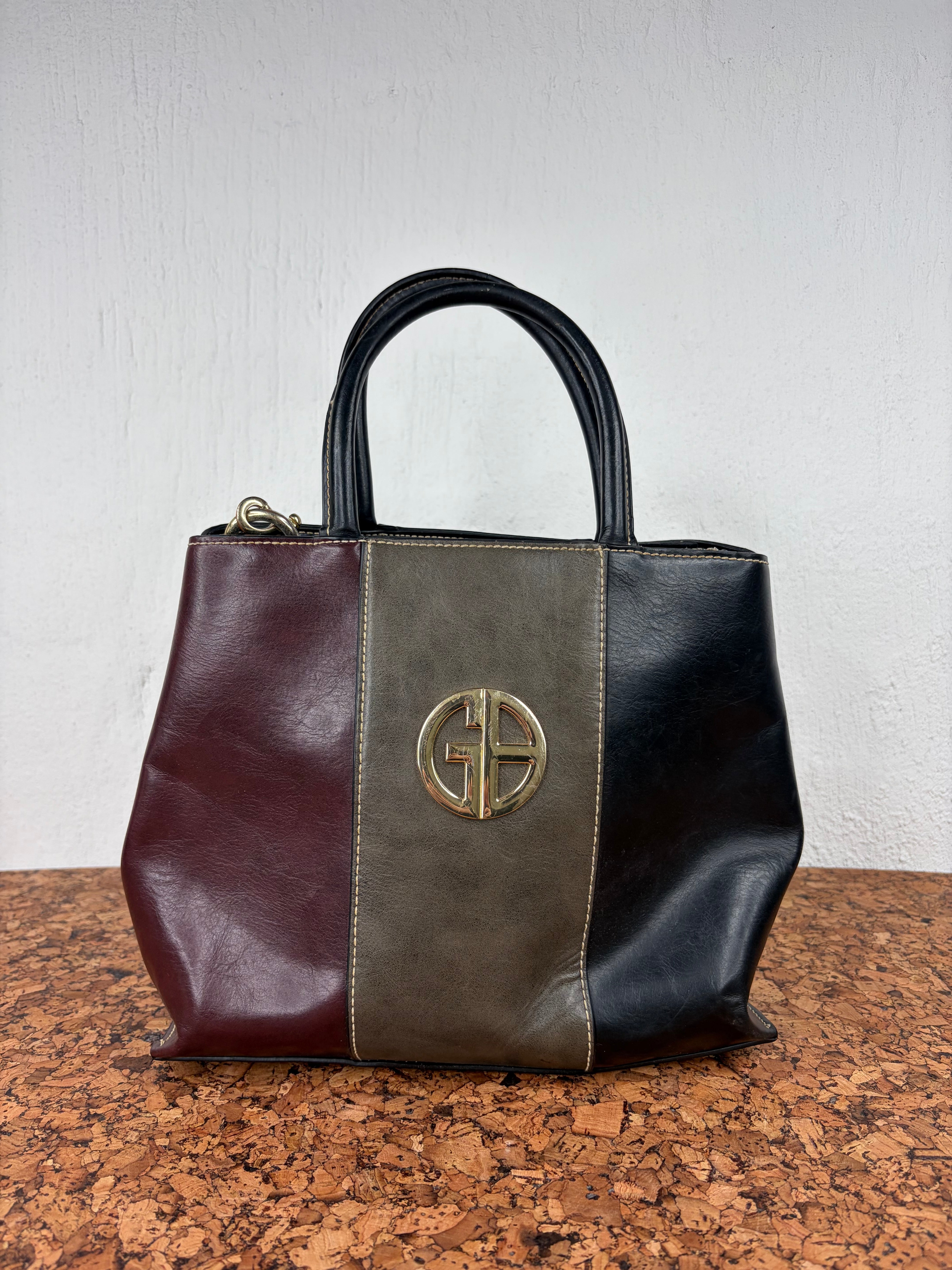 A leather handbag with a color-blocked design featuring burgundy on the left, olive green in the center, and black on the right, with a gold-tone circular cross logo on the front and black leather top handles.