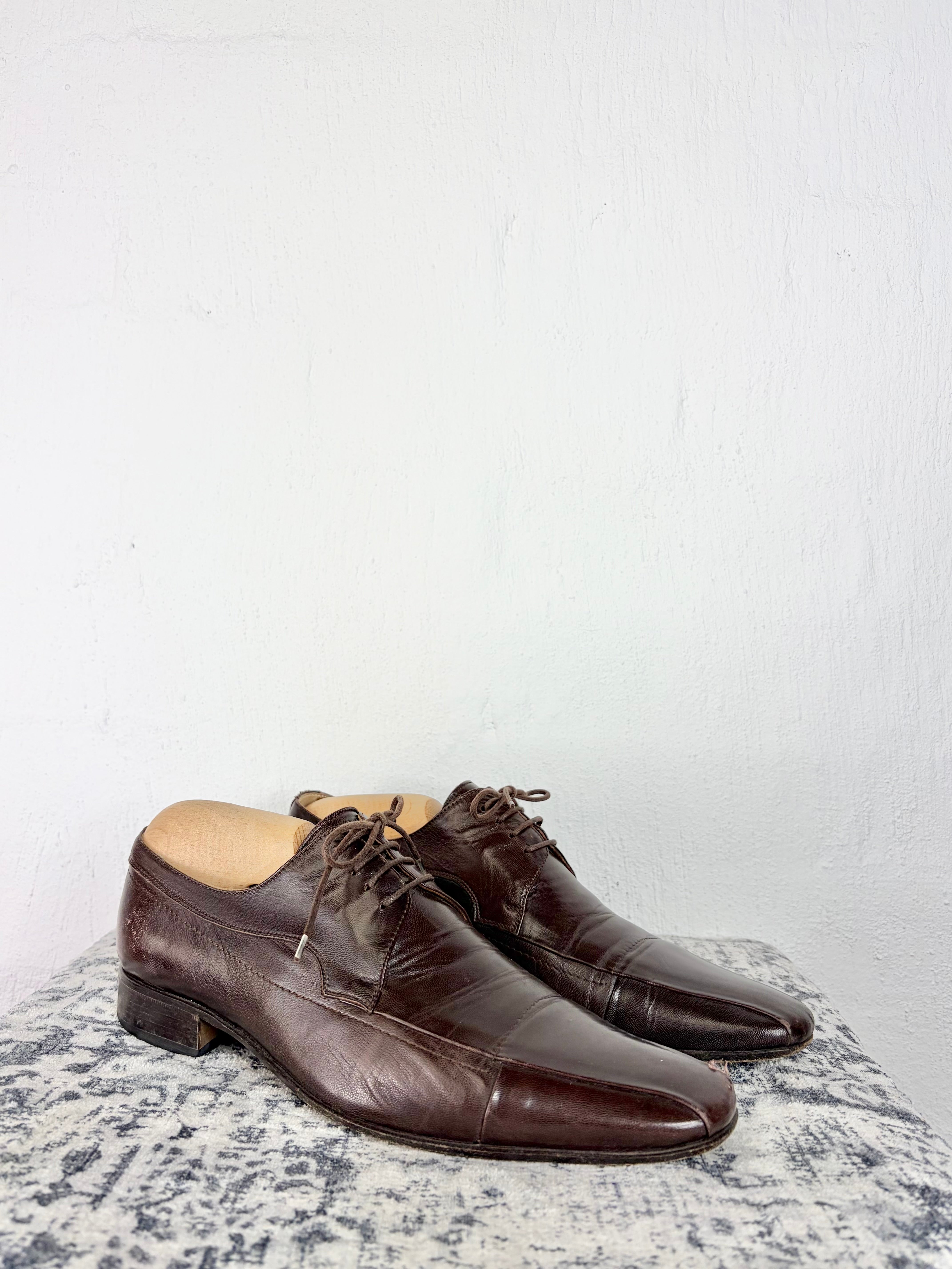 A pair of dark brown leather lace-up dress shoes with a cap toe, displayed on a textured gray and white surface against a plain white wall.