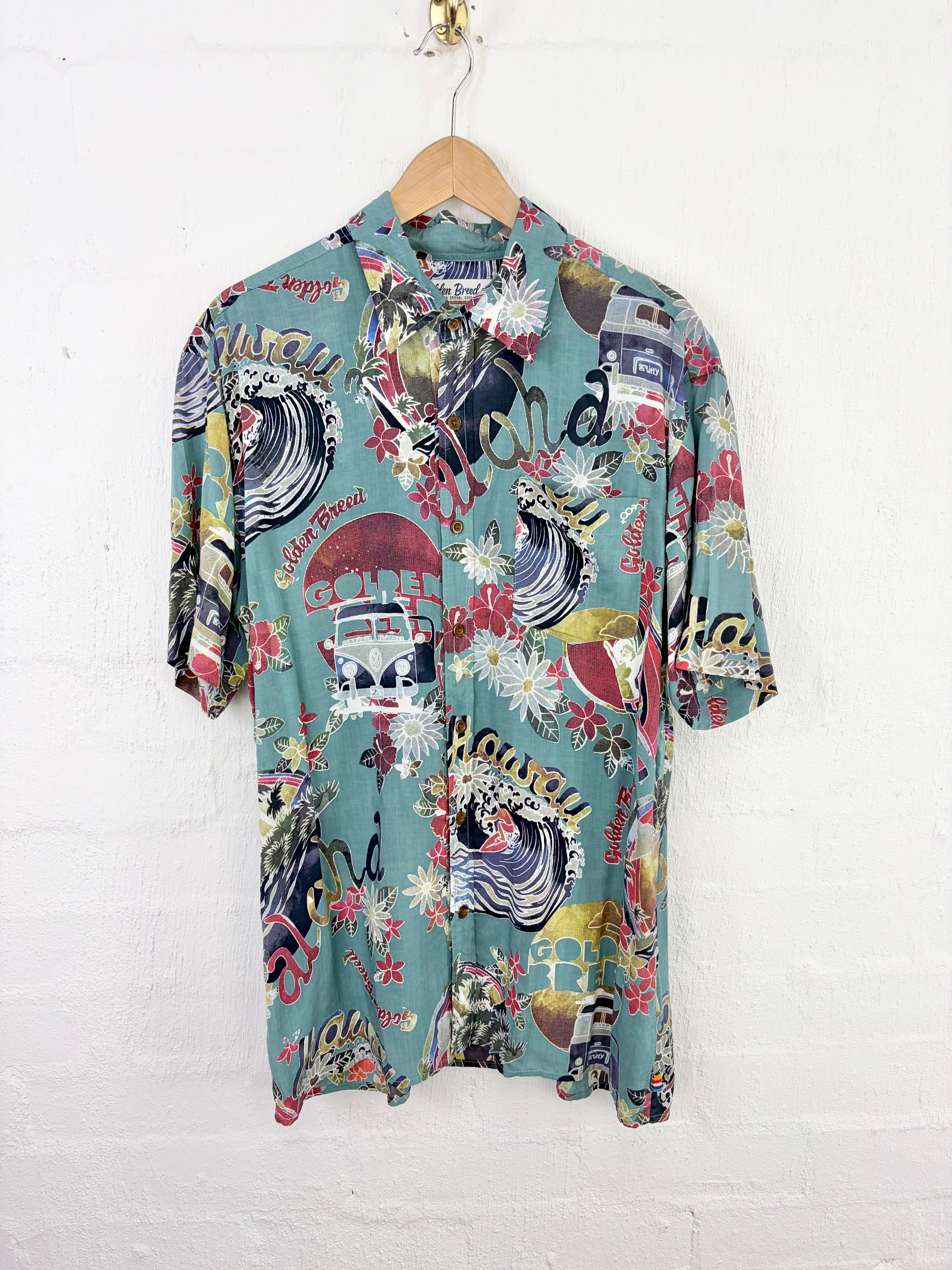 A short-sleeve button-up camp shirt with a colorful Hawaiian-themed print, hanging on a wooden hanger against a white painted brick wall.