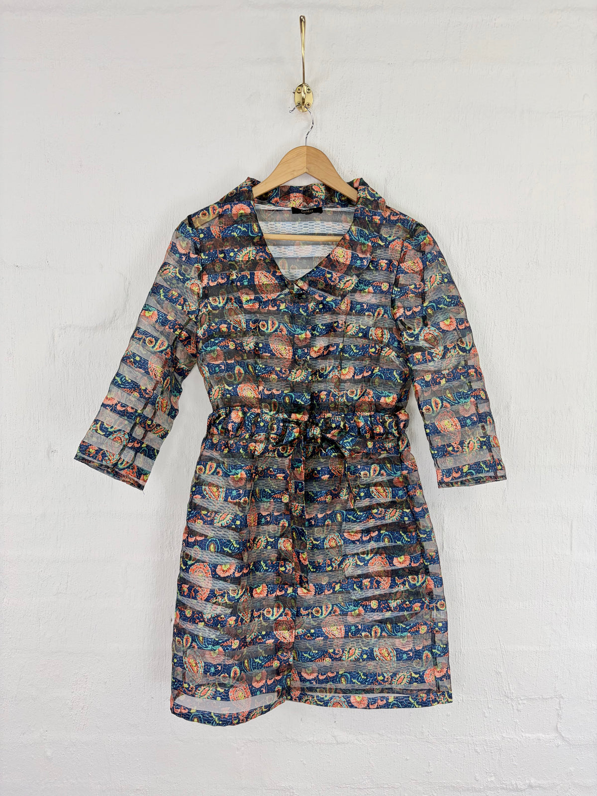 A long-sleeve wrap-style dress with a self-tie waist, featuring an all-over abstract pattern in navy, teal, orange, and beige, hanging on a wooden hanger against a white painted brick wall.
