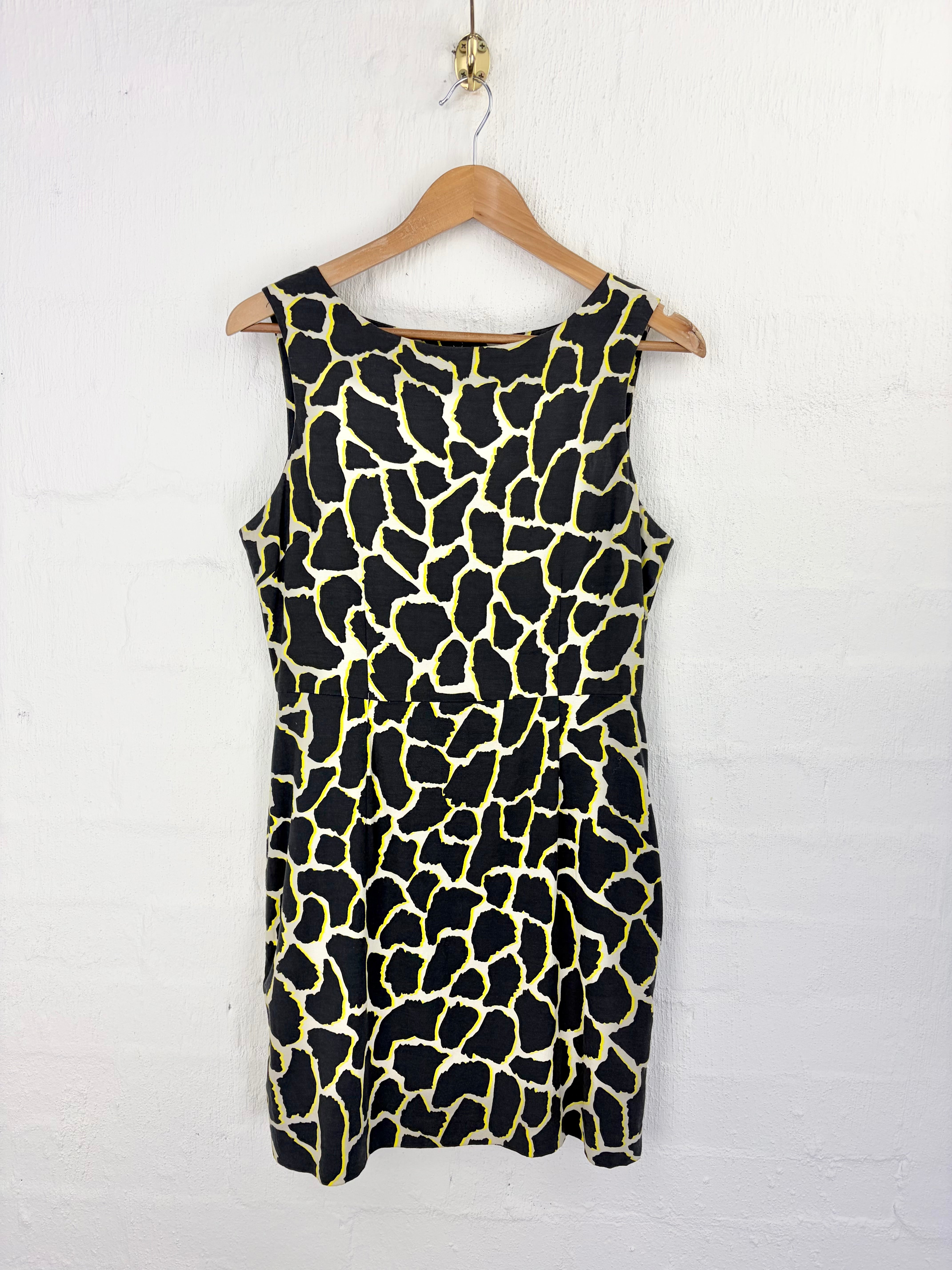 A sleeveless black dress with a yellow giraffe print pattern, hanging on a wooden hanger against a white painted brick wall.