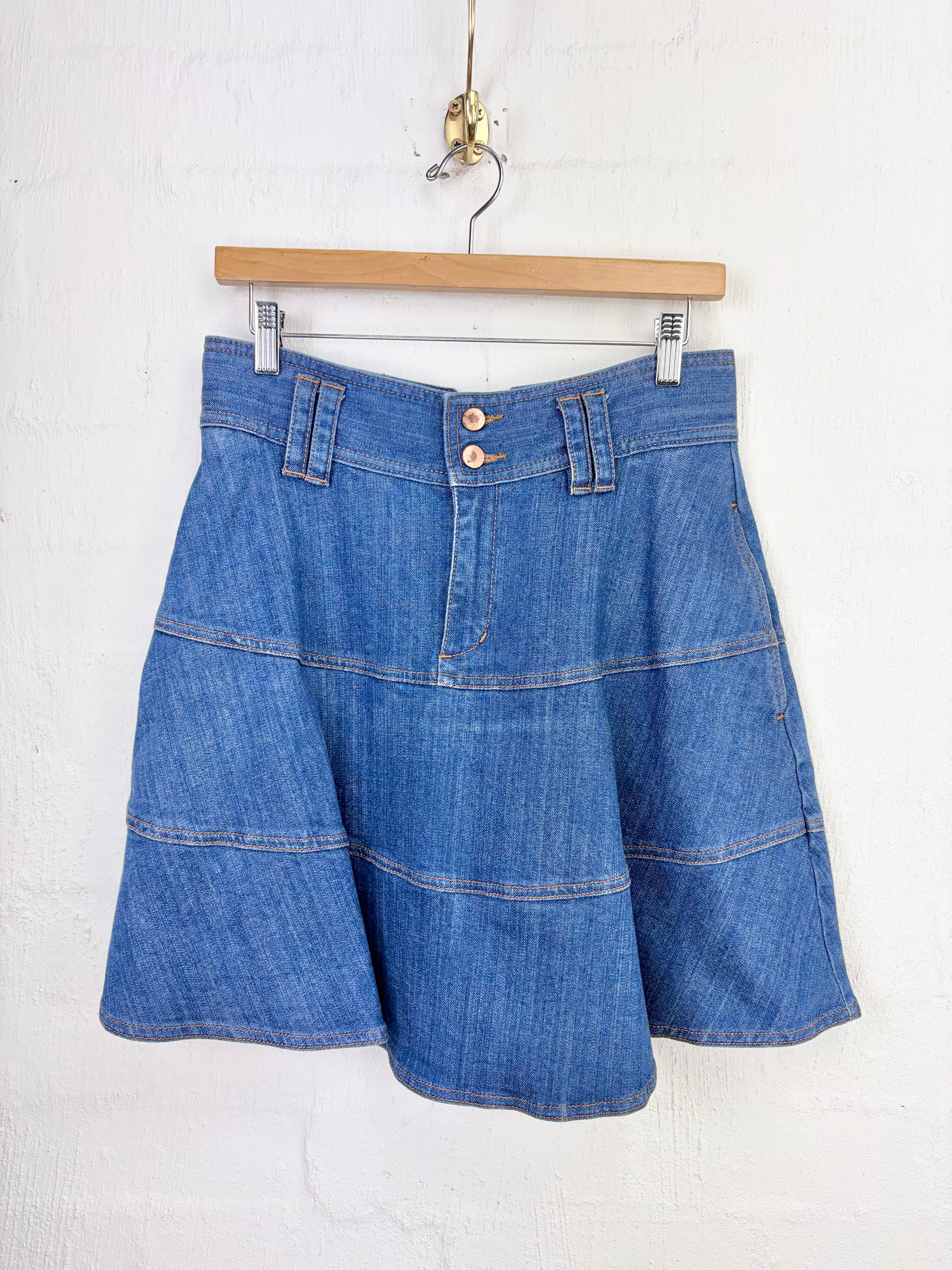A blue denim A-line mini skirt with horizontal seam detailing, hanging from a wooden hanger against a white textured wall.