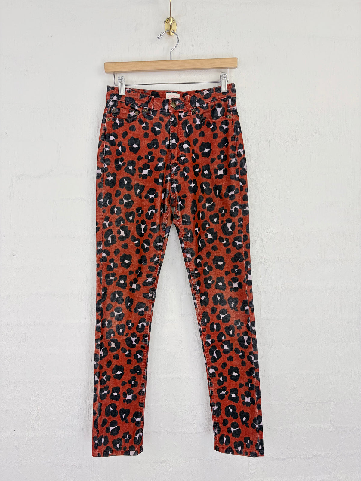Red leopard print pants hanging from a wooden hanger against a white brick wall.